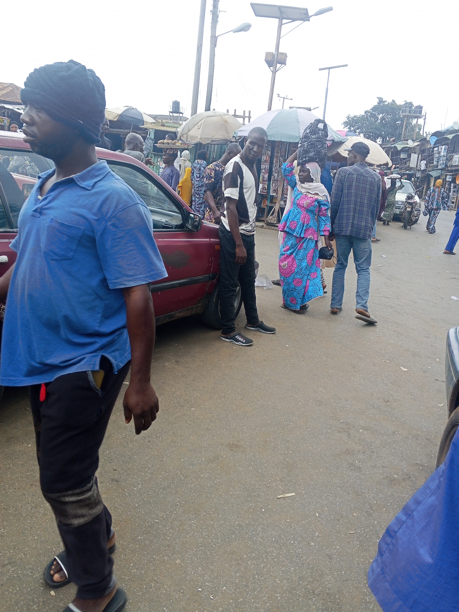 The image depicts a bustling street scene in Okene, Nigeria. A man in a blue polo shirt and black pants walks towards the foreground, his face turned to the left. Behind him, a maroon car is parked. Several other individuals are visible in the background, including a woman in a brightly colored blue and pink patterned dress, a man in a plaid shirt and jeans, and a person wearing a black and white striped shirt. The environment suggests a market or a busy commercial area, with stalls and umbrellas set up. A motorcycle is also visible. The ground is paved with asphalt.