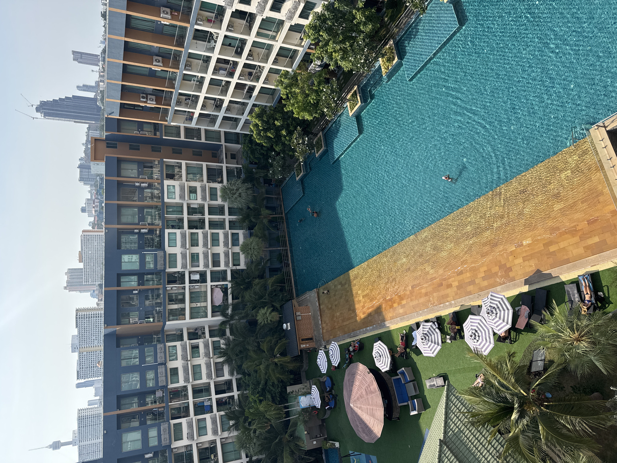 This elevated perspective showcases a modern residential or resort complex in Pattaya City, Thailand, under a clear, bright sky. The scene is dominated by a large, irregularly shaped swimming pool, tiled in vibrant turquoise, with a shallower section featuring warm yellowish-brown tiles. A few individuals are visible swimming or wading in the main pool.
The pool area is bordered by multi-story buildings characterized by contemporary architecture, featuring white, grey, and dark blue facades, numerous balconies, and external air conditioning units. Lush green trees, including several tall palm trees, are integrated into the landscaping around the pool and between the structures, adding a tropical ambiance.
Adjacent to the pool, a green leisure area, likely artificial turf, is furnished with dark sun loungers and several large, striped umbrellas (white and dark grey/black). Under and around these umbrellas, multiple people are seen relaxing, enjoying the outdoor setting.
In the background, a sprawling urban skyline unfolds, dense with numerous high-rise buildings and towering skyscrapers. A prominent construction crane indicates ongoing development in the city. Distinctly visible in the distant left, the Pattaya Tower landmark rises above the cityscape, firmly placing the location in Pattaya City. The strong, defined shadows cast by the buildings and umbrellas suggest the sun is high in the sky, indicating late morning or early afternoon. There is no legible text visible within the image.