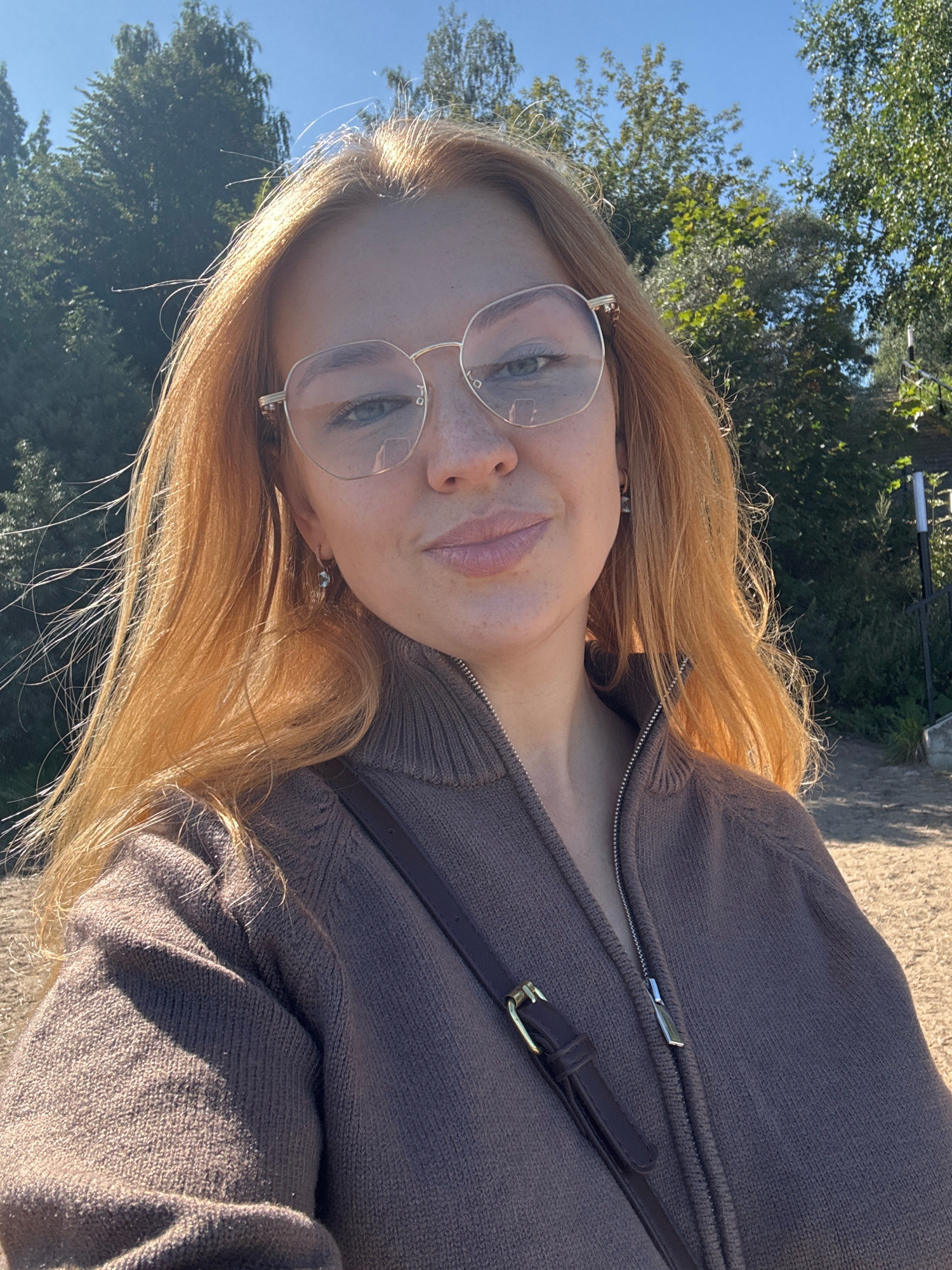 A young woman with blonde hair and glasses is taking a selfie outdoors. She is wearing a brown zip-up sweater. The background consists of trees and a clear blue sky. The location is the village of Ignatovo, Russia.