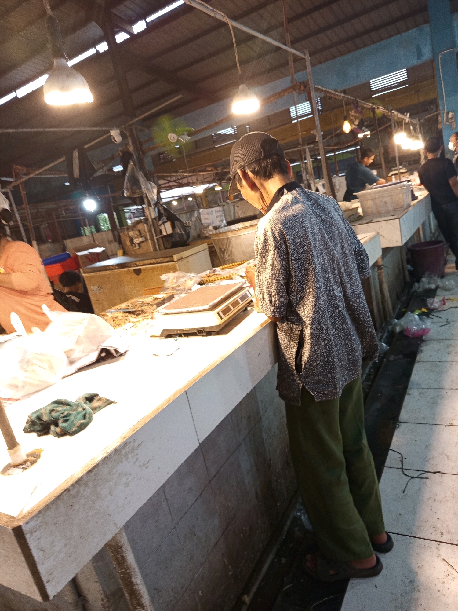 In the city of Karawang, Indonesia, a man wearing a cap, a patterned shirt, and green pants stands at a market stall. He is leaning over a counter that displays a weighing scale. To his left, there are plastic bags and a green cloth. In the background, other individuals are visible, as are market structures, lighting fixtures, and what appear to be food stalls. The environment suggests a busy, indoor market setting.