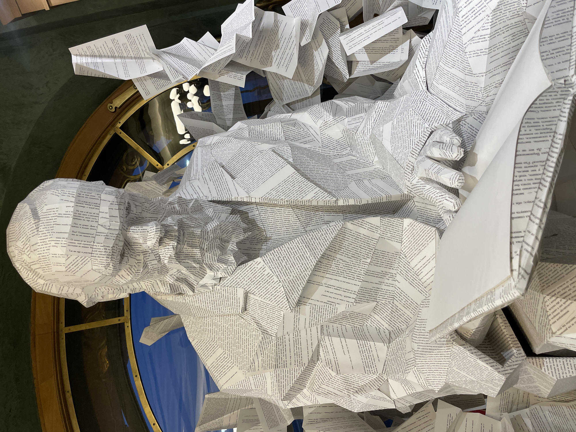 A three-quarter view captures a polygonal sculpture of a seated man, crafted entirely from white paper sheets printed with black Cyrillic text. The sculpture depicts a mature male figure with a beard, wearing a collared jacket, and looking downwards intently at an open, unprinted white book held in his hands. The paper is intricately folded and creased to form the geometric contours of the head, facial features, clothing, and hands. To the right and behind the sculpture, additional forms composed of crumpled and arranged text-printed paper are visible, suggesting an accumulation or part of the artistic installation. In the background, a large, ornate, arched window with a prominent golden frame reveals a clear blue sky with some scattered white clouds. The interior wall surrounding the window is a dark, textured green. This artistic installation is located within the **Russian National Library (Rossiyskaya Natsional'naya Biblioteka)** in Saint Petersburg, Russia.