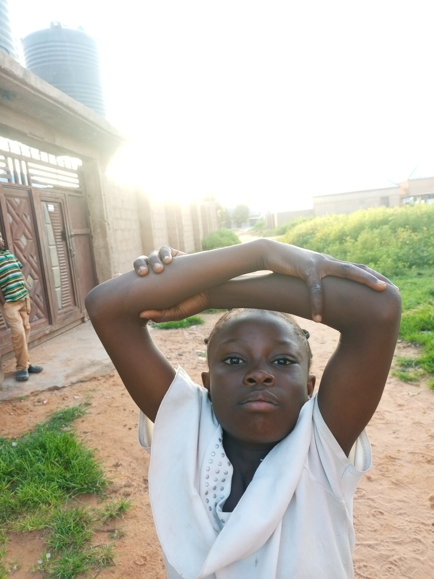 In an outdoor setting, likely in Tempure, Nigeria, a young Black child with braided hair is captured in a medium shot, looking directly at the camera. The child is wearing a light-colored garment. Their arms are raised, crossed over their head, with one hand resting on the elbow of the other arm. The background reveals a sandy ground with patches of green grass. To the left, a wooden gate with decorative metalwork is visible, and beyond that, a person wearing a striped shirt and light-colored pants stands. In the upper left corner, a large water tank sits atop a building. Further back, a wall constructed of cinder blocks and more vegetation can be seen. The sun is setting or rising, casting a bright light that overexposes parts of the scene.