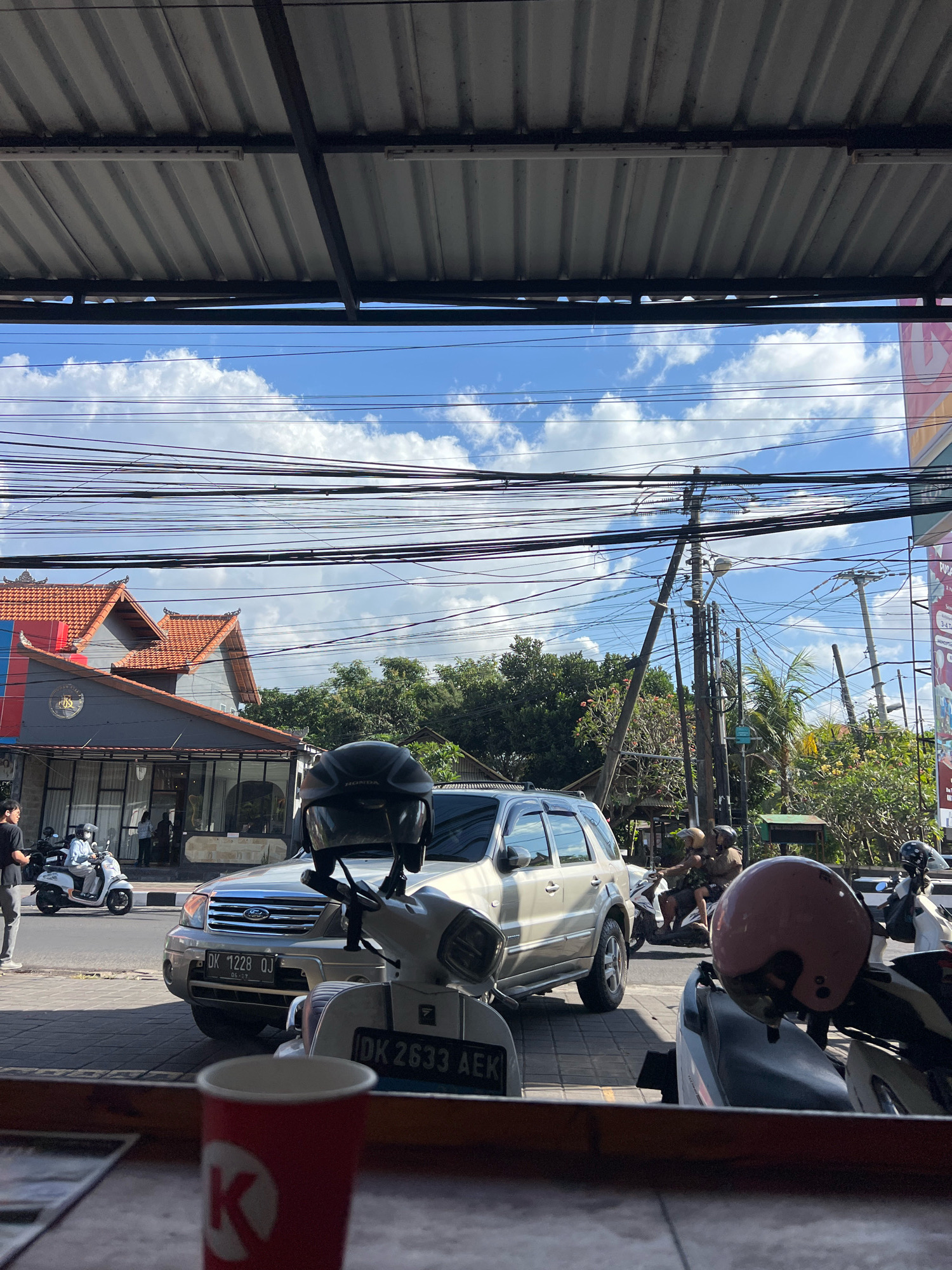 The image captures a street scene in Denpasar, Indonesia, under a bright blue sky with scattered clouds. A silver SUV, license plate DK 1228 OJ, is parked on the street. In front of the SUV, a white scooter with license plate DK 2633 AEK is visible, and another scooter is parked next to it. A person wearing a dark helmet is seated on the white scooter. To the left, a person in a black t-shirt is walking, and a white scooter with a person wearing a mask is in motion. The background features buildings with traditional Balinese architecture, including tiled roofs. Overhead, a dense network of power lines crisscrosses the sky, supported by wooden poles. The foreground shows a red cup with a white logo from what appears to be a coffee shop, and a portion of a table, suggesting the photo was taken from within a cafe or shop.