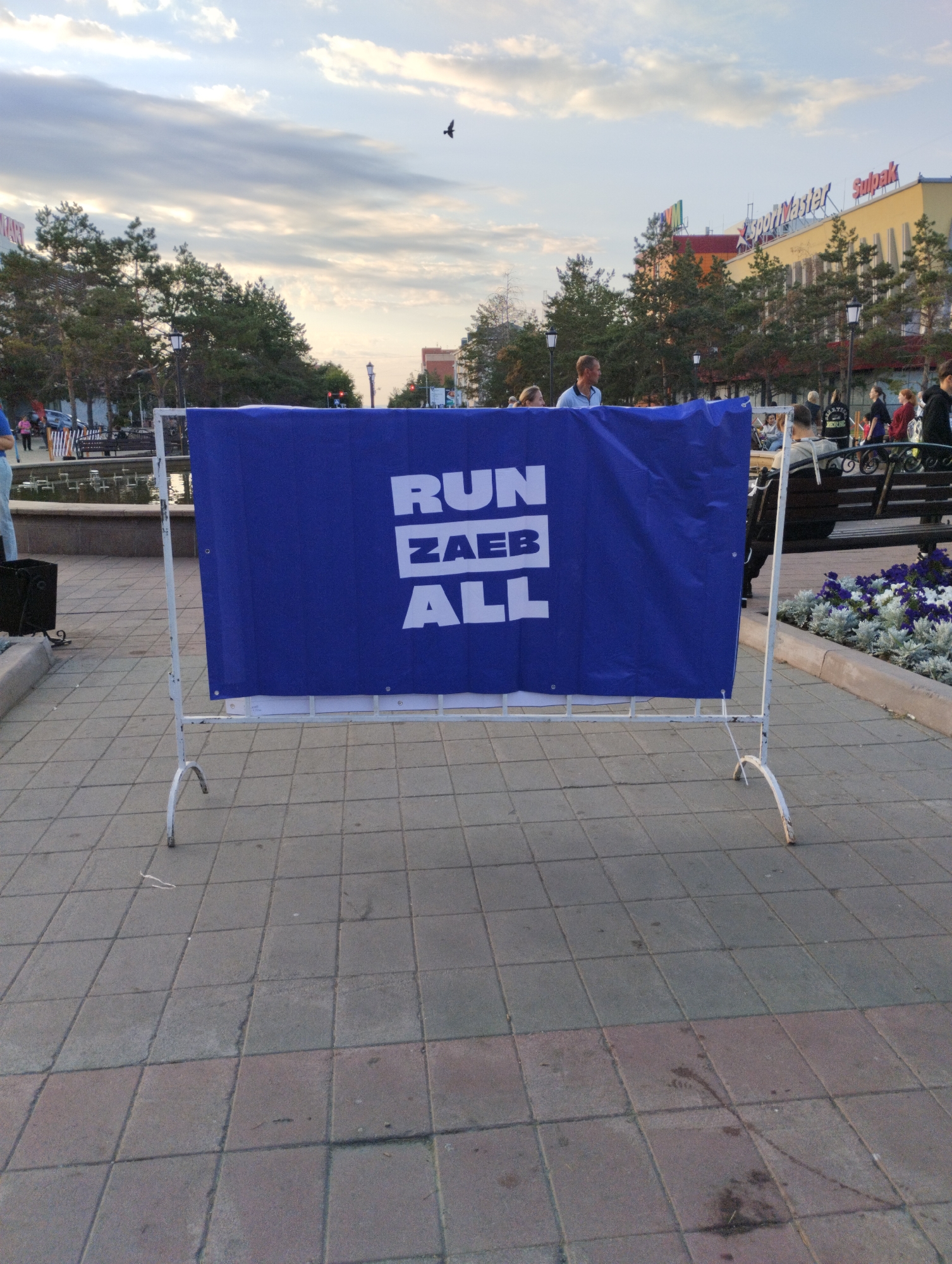 A banner with the text "RUN ZAEB ALL" in white capital letters on a blue background is displayed on a white metal stand in a city square. The banner is positioned in the foreground of the image. In the background, there are trees, a building with signs like "SportMaster" and "Sulpak," and people walking or sitting. The sky above is cloudy, with a bird in flight. The ground is paved with square tiles. The scene appears to be an outdoor event or gathering.