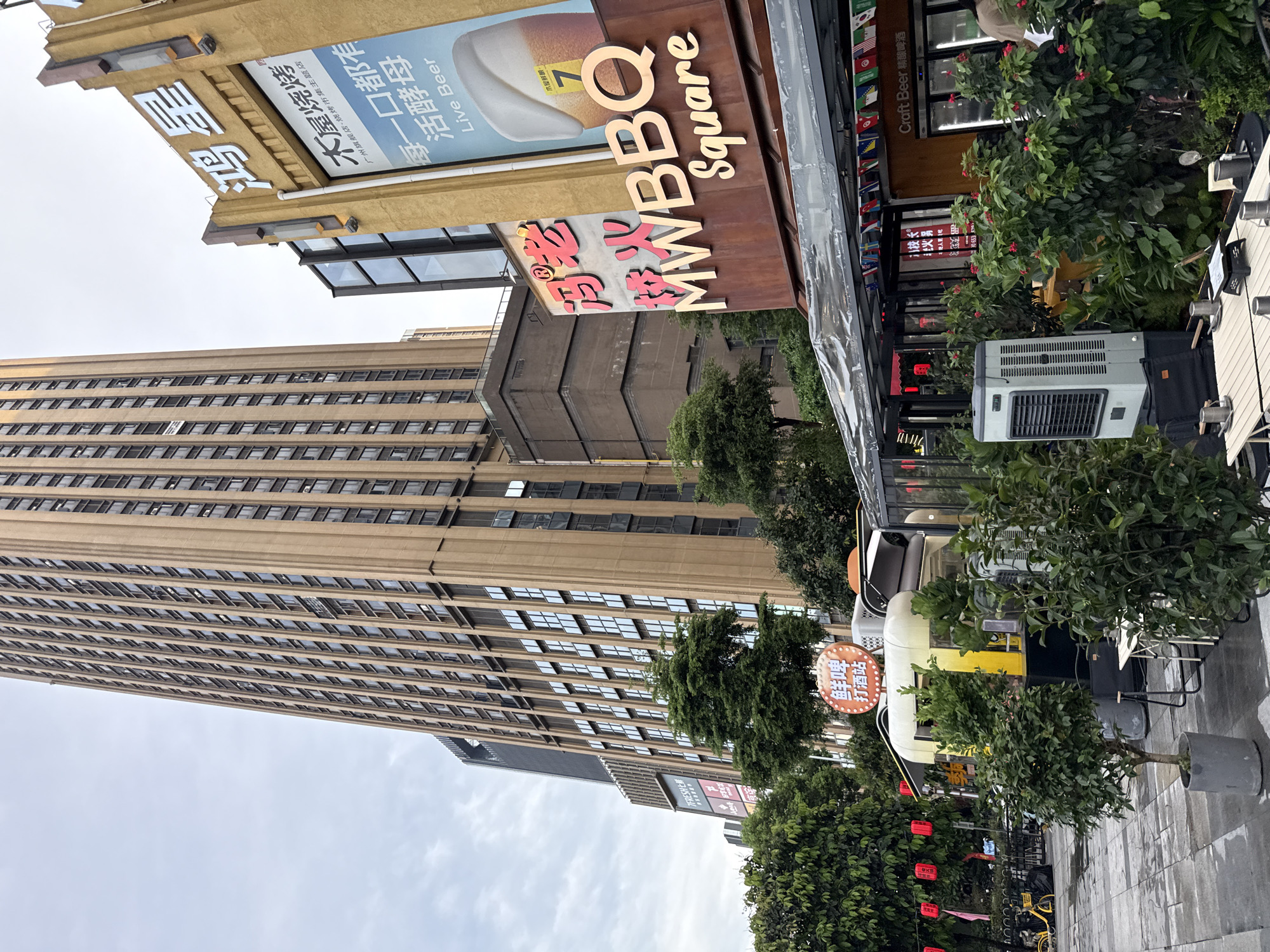 This is a vertical, outdoor, eye-level shot of a commercial street scene in Haizhu District, China. The sky is overcast and bright, suggesting a cloudy day, possibly in the morning or afternoon. The dominant subjects are buildings, signage, and greenery.
In the foreground, a portion of an outdoor seating area with tables and chairs is visible, partially covered by a clear plastic awning. A light-colored outdoor air conditioning unit is prominent. To the right of the seating area are small, potted trees and lush green bushes. Red lanterns are strung up on a fence in the middle ground.
Behind the outdoor seating, a building with a dark brown facade features signage that reads "MW BBQ Square" in English and "木屋烧烤" (MuWu ShaoKao - Wooden House Barbecue) in Chinese. Above this, another sign advertises "Craft Beer 精酿啤酒" (JingNiang PiJiu - Craft Beer) with flags from various countries displayed. Further to the left, a tall, beige, multi-story building dominates the background, its facade covered in rows of windows. Next to it, a darker, Brutalist-style building is partially visible.
Several pieces of text are legible in the image, including restaurant names, advertising slogans like "每一口都有活酵母 Live Beer" (Every Mouthful Has Live Yeast Live Beer), and street vendor signage like "鲜啤 打酒站" (XianPi DaJiuZhan - Fresh Beer Takeaway Station). There are no people clearly visible in the frame, nor any specific activities taking place other than the implied operation of businesses. The overall impression is one of a bustling urban commercial area with a focus on food and beverage.