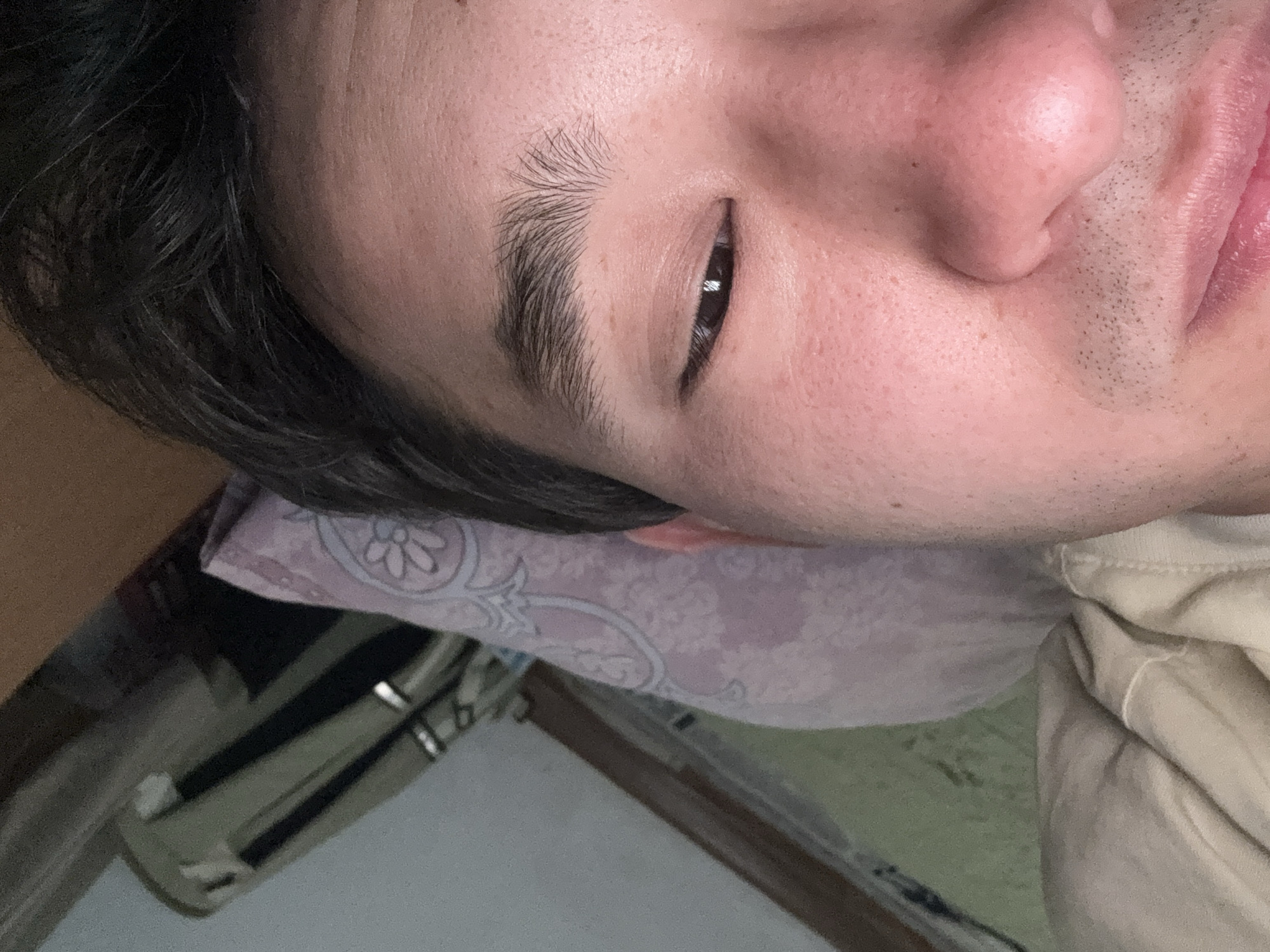 A close-up, eye-level, indoor shot shows a person's face from the nose up, with the left side of the face being most prominent. The person has brown hair, dark eyebrows, and dark eyes. The person is wearing a light-colored hooded sweatshirt. The background shows a pillow with a floral pattern, a portion of a bed, and what appears to be a wall with some objects attached to it. The lighting is dim, creating shadows on the face.