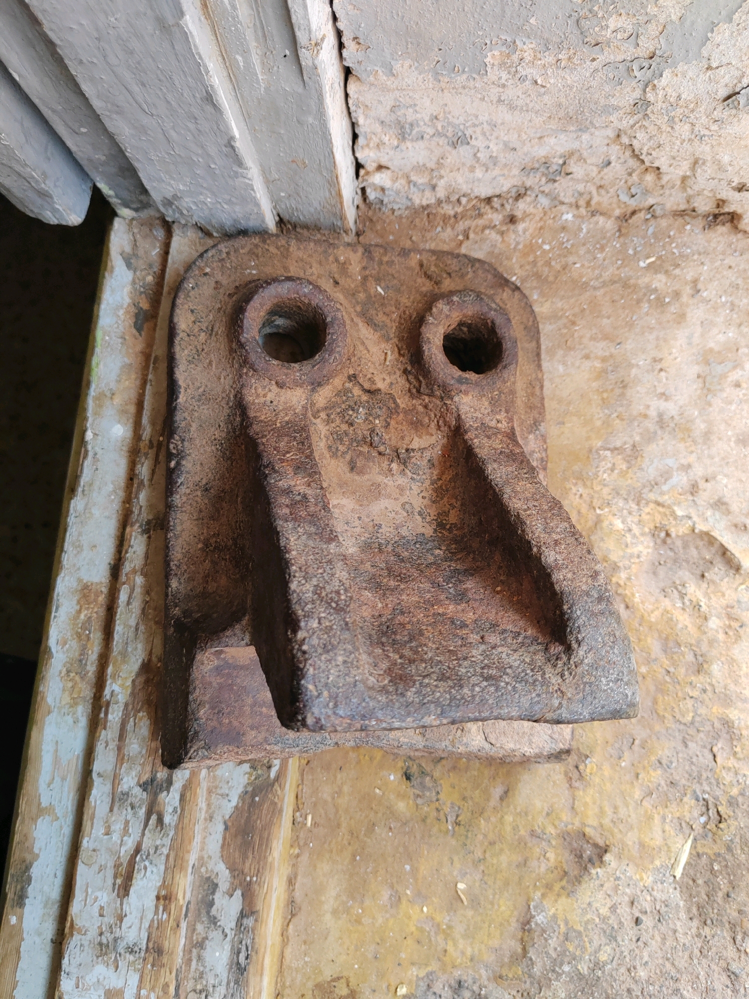 The image shows a close-up, high-angle shot of a rusted metal object attached to a rough, textured wall. The metal object is rectangular with two circular holes near the top, resembling eyes, and two angled protrusions below them, giving it a face-like appearance. It is heavily corroded and has a brown, rough texture. The wall behind it is uneven and appears to be made of concrete or plaster, with patches of peeling paint or discoloration in yellow and grey hues. To the left of the metal object, there is a wooden structure with peeling white paint, possibly a door frame or window sill. The scene is dimly lit, and the overall impression is one of neglect and age. The location is identified as Mougheul, Algeria.