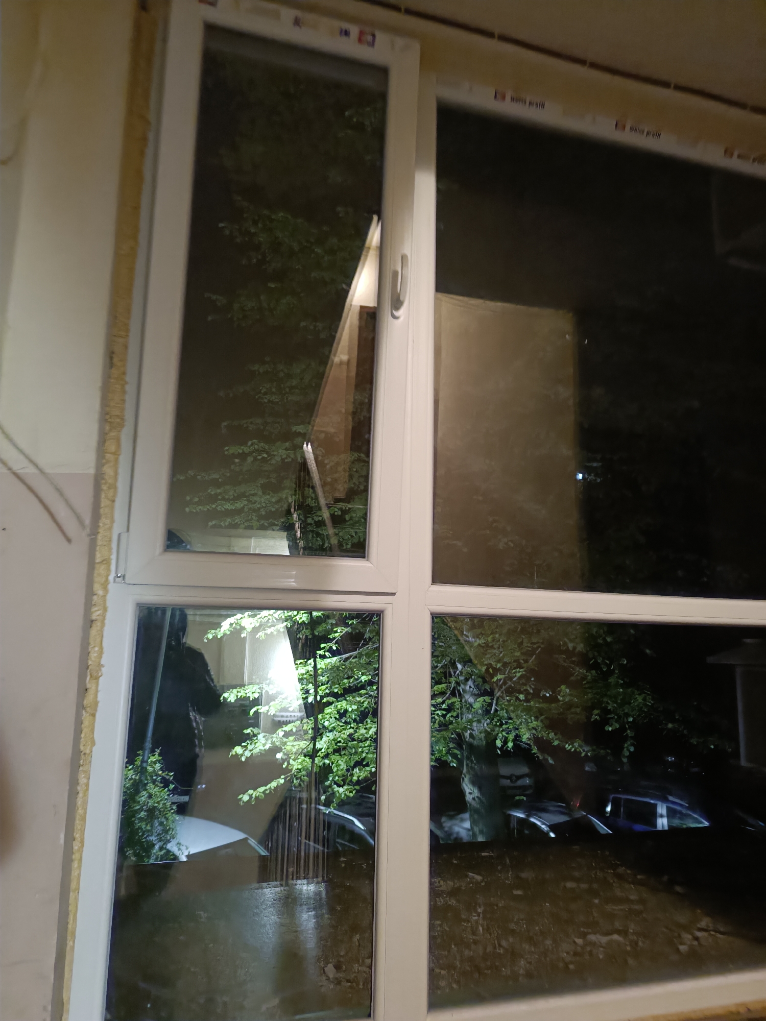The image depicts a view from inside a building, looking out through a partially open window. The window is white-framed and divided into four panes. It is nighttime, and the scene outside appears to be a street with trees and parked cars. There is a streetlamp illuminating a nearby building and foliage, visible in the lower left pane. The overall environment suggests a residential area, and it is raining or has recently rained, as evidenced by the wet pavement. While the precise location in Pleven, Bulgaria, cannot be determined with certainty from the image, the architectural style and vegetation are consistent with the city's environment.