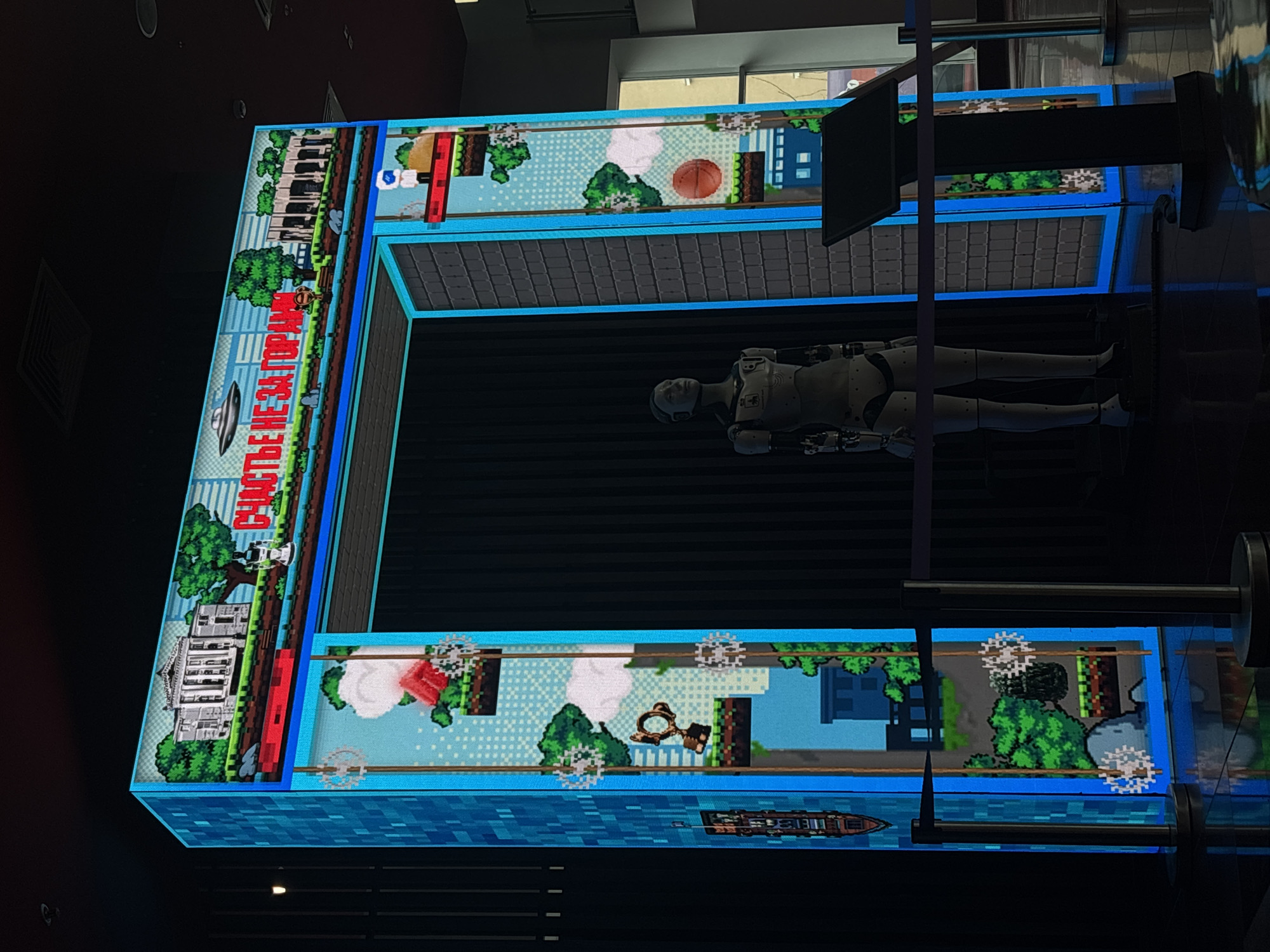 An articulated humanoid robot, light-colored, lies supine within a large, C-shaped digital display in an indoor setting, likely an exhibition or museum in Perm, Russia. The robot occupies the central dark space of the installation, its form reflecting off the dark surface below.
The surrounding display features vibrant pixel art graphics, creating a continuously scrolling landscape reminiscent of retro video games. The scenes depicted vary across the frame:
* On the left side, a detailed pixelated white building, resembling a classical mansion, is surrounded by green trees, with a red train nearby. Above this, a flying saucer (UFO) is visible. Bold red text, partially obscured, reads "СЧАСТЬЕ НЕ ЗА ГОРАМИ" (Happiness is not far off / Happiness is just around the corner) in Cyrillic script.
* The top section of the display shows more trees, buildings, and a basketball.
* The bottom section features a large red pixelated rose or flower, a key, and a ship sailing on blue pixelated water. Small, spiky circular objects are scattered across this part of the display.
The overall aesthetic combines a futuristic robotic element with nostalgic pixel art, creating a thought-provoking display. The lighting is dim, highlighting the illuminated screen and the robot.