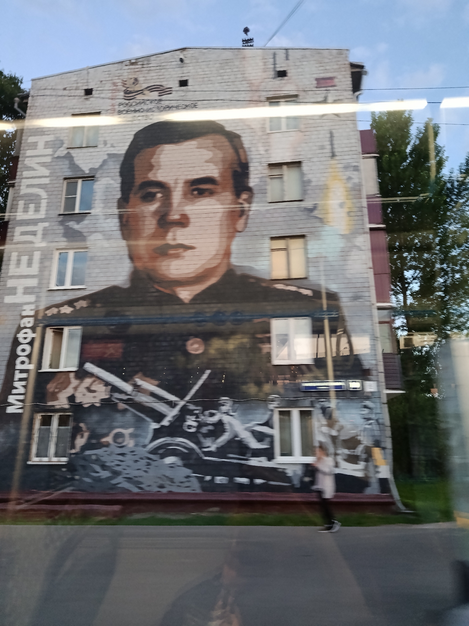 The image shows a mural painted on the side of a building. The mural depicts a portrait of a man in military uniform above an image of an artillery crew operating a cannon. To the left of the mural, vertically oriented, is text in Cyrillic that reads "Митрофан Неделин". The building is gray brick and has multiple windows.
The building is located on Beregovoy Proezd in Moscow, Russia. The photo was likely taken from inside a vehicle. A blurred figure can be seen walking in front of the building. Trees are visible to the right of the building.