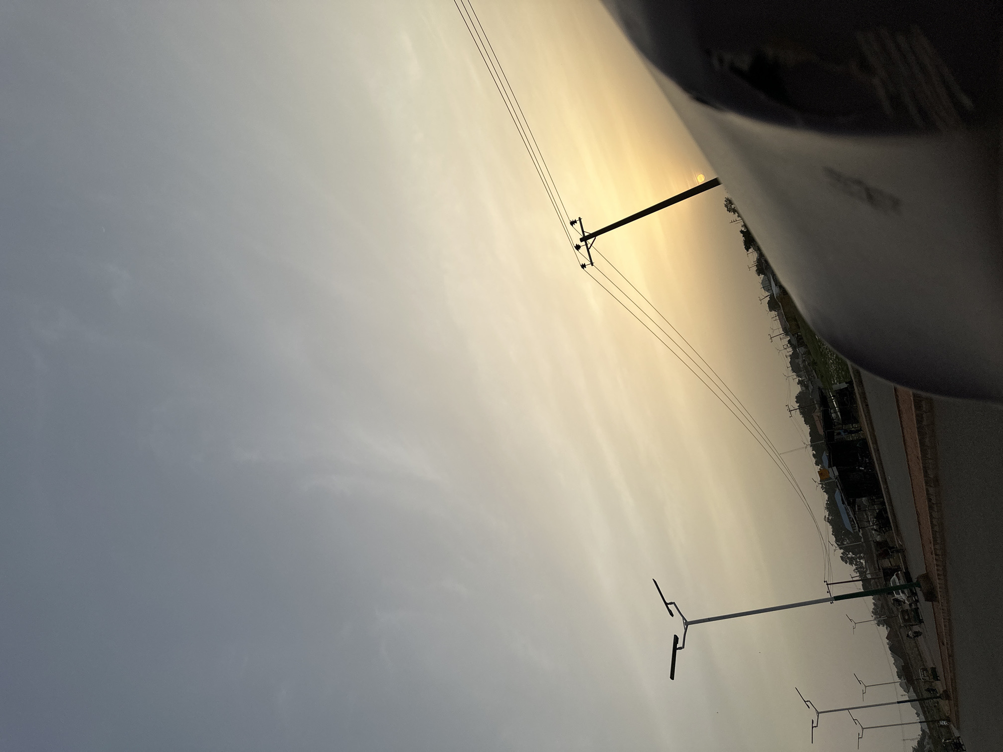 The image captures an outdoor scene at either sunrise or sunset, characterized by a dramatic sky and roadside infrastructure. The sky transitions from a soft, muted grey on the left to a warm, luminous golden-yellow and orange on the right, where the sun is a bright, distinct orb, partially obscured by a dark foreground element. Faint, wispy cloud formations are visible across the sky.
A prominent electricity pole stands silhouetted against the bright portion of the sky, with multiple power lines extending horizontally across the frame. Further down, several modern-looking streetlights with distinctive 'T' or 'Y' shaped fixtures are positioned along the edge of a paved road. The road runs along the lower right side of the image, appearing to stretch into the distance.
Along the roadside, indistinct buildings, possibly houses or small shops, are visible, along with scattered trees and vegetation, mostly silhouetted against the lighter sky. A large, dark, curved object occupies the right side of the foreground, suggesting the image was taken from within a vehicle, looking out a window. No people or specific activities are discernible in the frame. The golden hour lighting creates a serene and somewhat hazy atmosphere. There is no clearly legible text visible in the image.