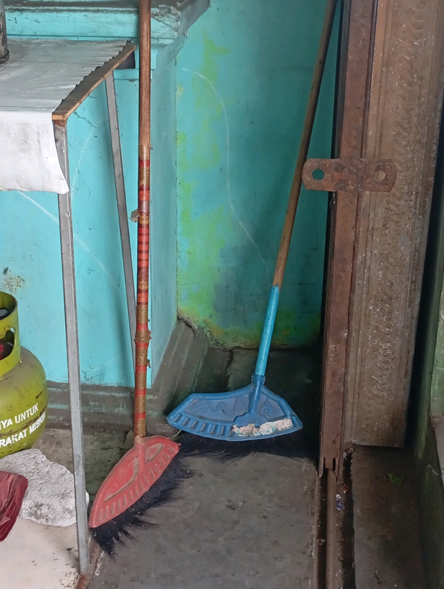 The image shows two brooms leaning against a blue and green mottled wall in what appears to be an outdoor or semi-enclosed space, possibly a back alley or storage area. The broom on the left has a red handle with a textured pattern and black bristles. It rests on the ground next to a yellow gas cylinder with white lettering. To the right of this broom, a second broom with a light blue handle and blue bristles is leaning against a dark brown, possibly wooden or metal, frame. The ground is concrete and appears wet in places. The wall behind the brooms is painted in a weathered, uneven style, with streaks of different colors. The overall scene suggests a utilitarian or storage area, likely in a residential or commercial setting in Babelan, Indonesia.