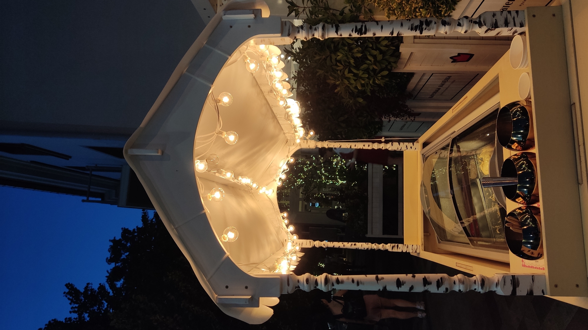In Krasnodar, Russia, at dusk, a white ice cream cart is displayed outdoors. The cart features a quaint, house-like roof adorned with a string of warm, glowing Edison bulbs. The support posts of the cart are designed to resemble birch trees with black markings. Inside the cart, a glass display case shows containers of what appears to be ice cream. In the foreground, three shiny silver bowls hold what seem to be utensils. Two small white paper cups are also visible on the cart's surface. In the background, blurred figures of people can be seen, and lush green foliage surrounds the cart. The sky above is a deep, twilight blue.