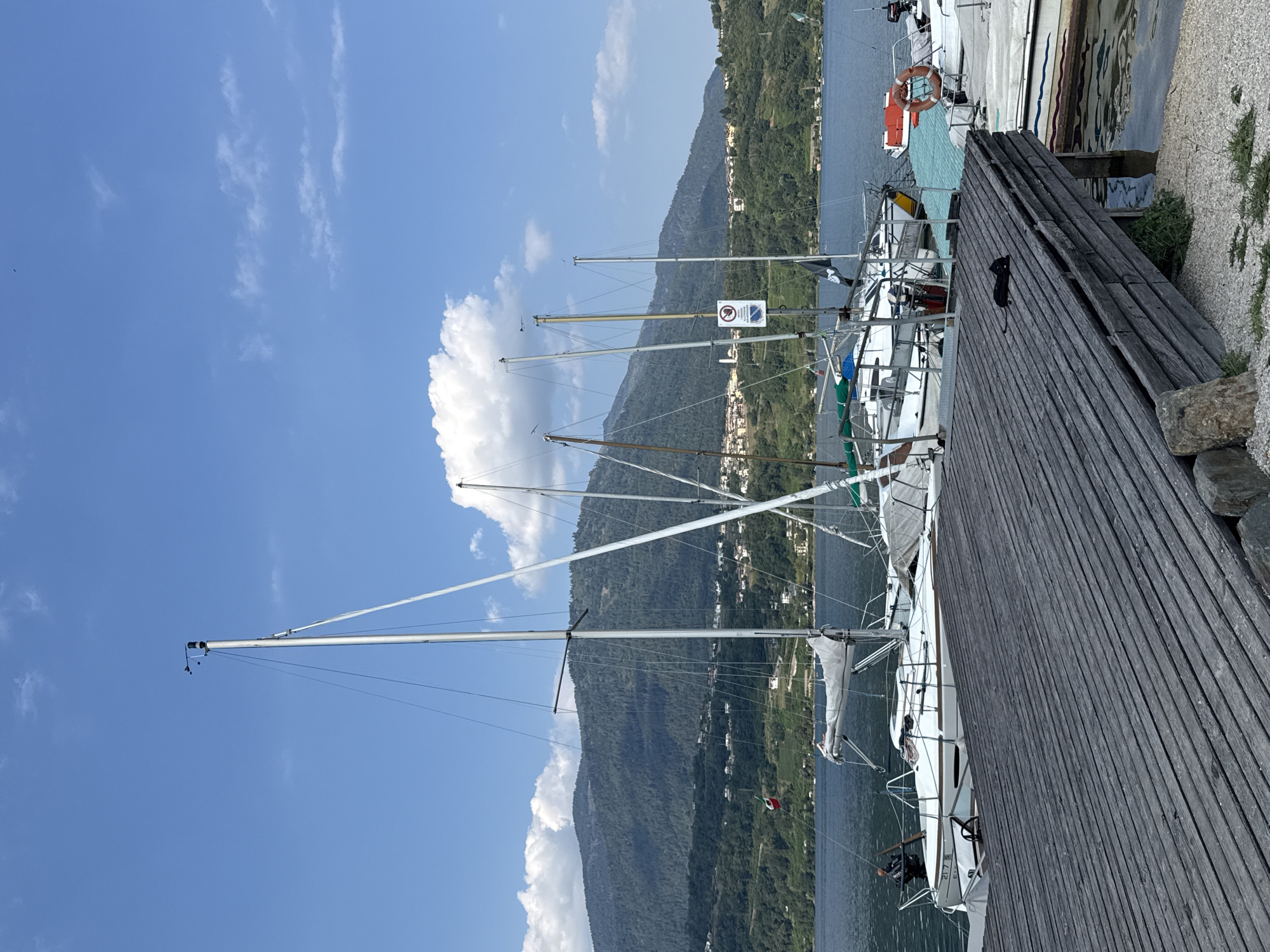 The image depicts a sunlit scene at a marina in Calceranica al Lago, Italy. A wooden dock, constructed from weathered grey planks, occupies the foreground, angled to the right. Several sailboats are moored alongside the dock, their white hulls and tall masts dominating the middle ground. The lake water is a deep blue, reflecting the clear sky. In the background, a verdant, tree-covered mountain range rises, dotted with small buildings, suggesting a town nestled on its slopes. The sky is a bright blue with scattered white clouds. A black object, possibly a sleeping cat, is positioned on the wooden dock in the lower right quadrant of the image. A life preserver ring is visible on one of the boats. The overall impression is one of a peaceful, picturesque day at the lake.