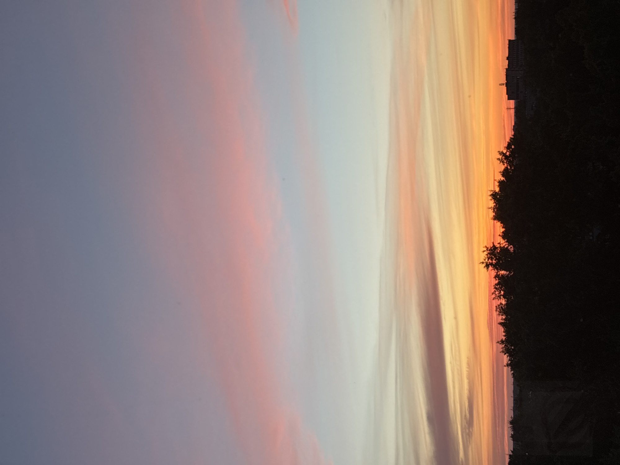 The image depicts a sunset over a city skyline. The sky is a gradient of colors, starting with a deep blue at the top, transitioning through pink and light blue, and ending with vibrant orange and yellow hues near the horizon. Wispy clouds are present in the upper portion of the sky, reflecting the pink tones. Below the sky, a dark silhouette of trees spans across the lower third of the image. In the far distance, a building complex with lights is visible, appearing as a dark shape against the brighter horizon. The overall scene is peaceful and atmospheric, characterized by the dramatic colors of the sunset.
