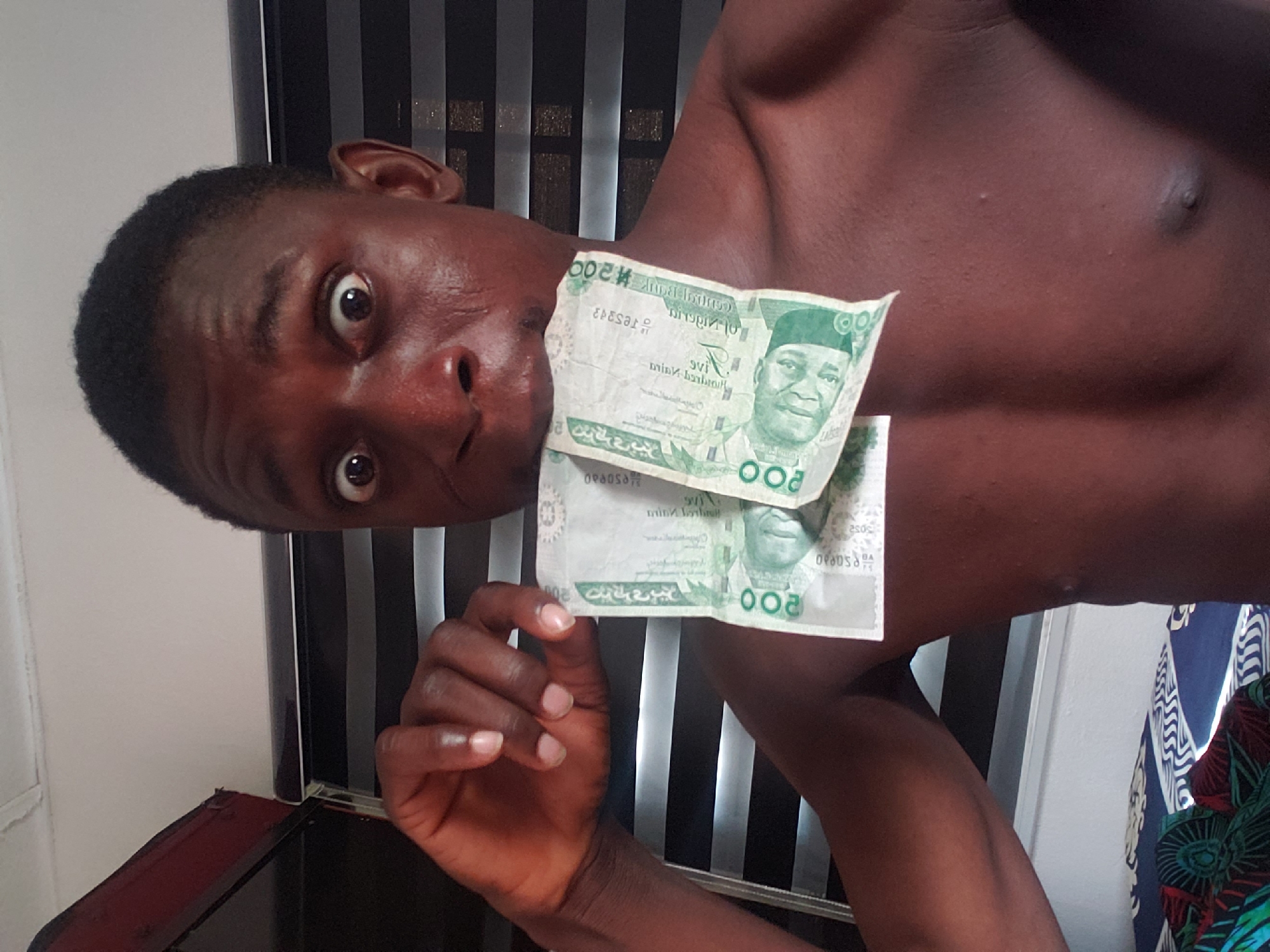 The image captures a young Black man posing indoors, likely in Adonkolo, Nigeria. His head is tilted sharply to the left, and he looks directly at the camera with wide, expressive eyes, conveying a sense of surprise, shock, or humorous exaggeration. He is shirtless, revealing his torso.
He holds two green Nigerian Naira banknotes, each with a denomination of 200, in front of his mouth and chin with his right hand. The banknotes feature the portrait of Ahmadu Bello. Visible text on the currency includes "CENTRAL BANK OF NIGERIA," the numeral "200," and partial serial numbers such as "082050" and "3ES8942."
The background on the left consists of a plain white wall. Behind his right shoulder and extending downwards, a dark object with distinct vertical black and silver/grey stripes is visible, possibly furniture or wall decor. On the far right, a piece of patterned fabric with blue, white, and green designs, likely clothing or a curtain, adds a touch of color to the scene. The lighting suggests it is daytime. The man's pose and exaggerated expression, coupled with the display of currency, indicate a playful or meme-inspired interaction with money.