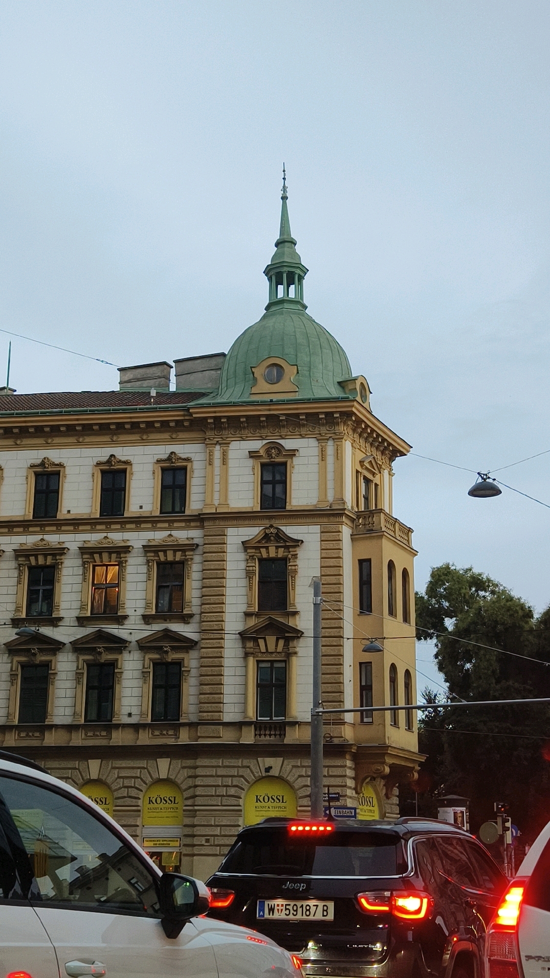 The image shows a street scene in Vienna, Austria, focusing on a corner building with a green dome. The building has a classical architectural style with white and yellow facade elements, ornate window frames, and decorative moldings. A tall, slender spire tops the green dome. In the foreground, several cars are visible, suggesting traffic on a city street. One car is black with a "Jeep" logo and an Austrian license plate that reads "W 59187 B." Another white car is partially visible in the lower left corner, and a white car's taillight is seen on the right. Yellow signs with the text "KÖSSL" and "KUNST & TEPPICH" are displayed on the lower level of the building, indicating a shop or business. Overhead, streetlights and power lines are present. The sky is overcast and appears to be either early morning or late afternoon. The overall impression is a snapshot of urban life in Vienna.