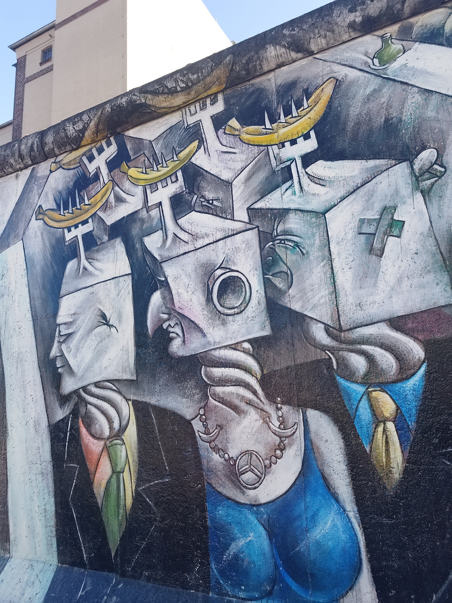 This is a photograph of a section of the Berlin Wall in Berlin, Germany. The painting depicts three figures, each with a cube-shaped head. The cube-heads are different shapes and styles. Each of the figures appears to be wearing some sort of clothing. The heads are surmounted by yellow bananas with spikes. The overall style is surreal and symbolic. The mural is weathered, with cracks and signs of age. The painting is on the Berlin wall. The building is visible in the background.