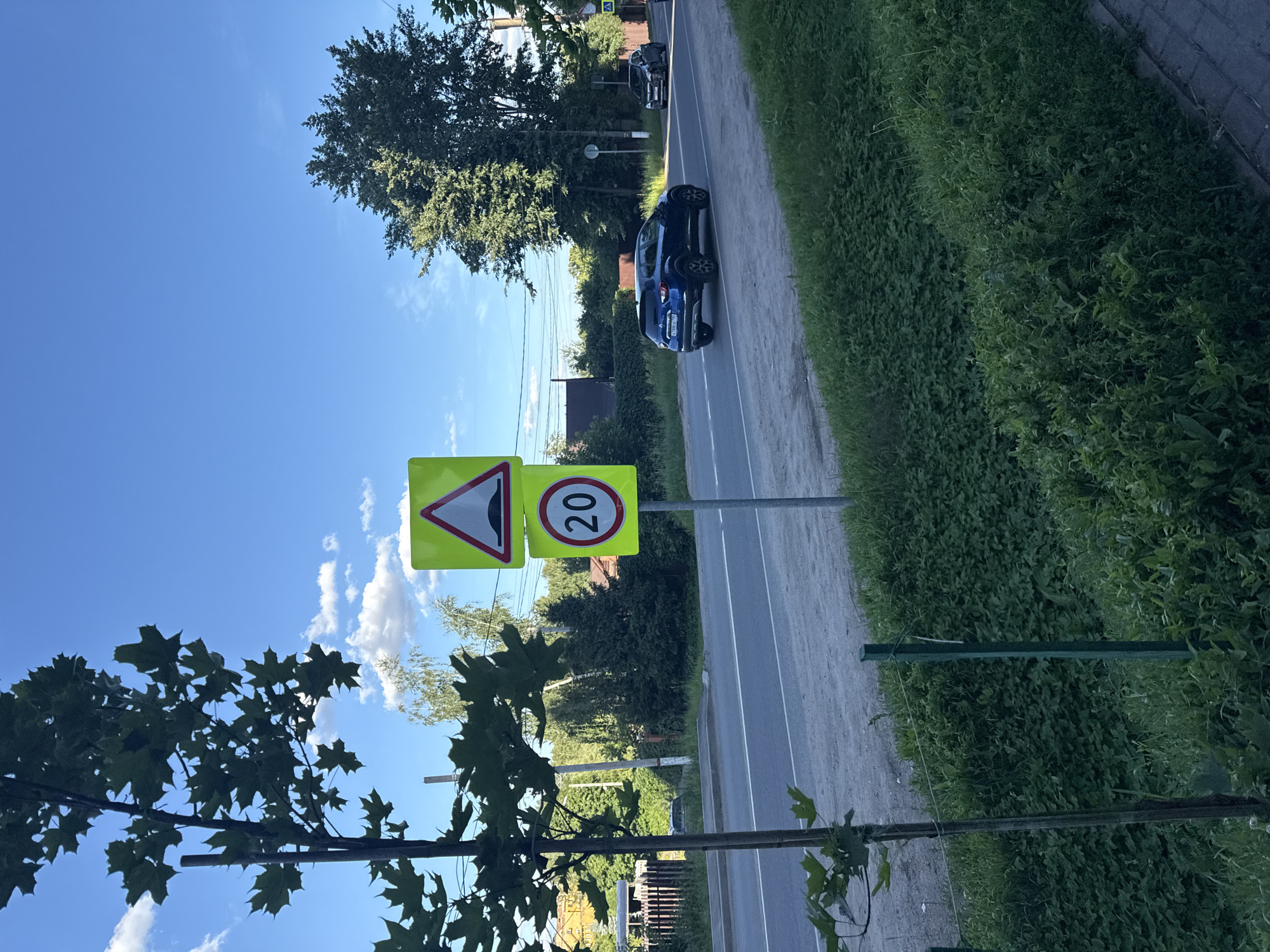 The image depicts a roadside scene in Golitsyno, Russia, under a clear blue sky with scattered white clouds. Two prominent yellow road signs are mounted on a single pole on the right side of the frame. The upper sign is a warning triangle with a black silhouette of a speed bump, indicating an upcoming speed bump. The lower sign is circular, with a red border and the number "20" in black, signifying a speed limit of 20 kilometers per hour.
A dark blue SUV is driving on the asphalt road, moving away from the viewer and towards the right. To the right of the SUV, another dark-colored car is visible further down the road. The road has white lane markings.
On the left side of the road, a young tree with green leaves is growing. Behind the tree and the road, there is lush green vegetation, including a grassy embankment and dense bushes. In the background, several houses with red and brown roofs are visible, surrounded by trees and power lines. The foreground shows a paved sidewalk and more greenery. The overall impression is a suburban or rural road on a sunny day.