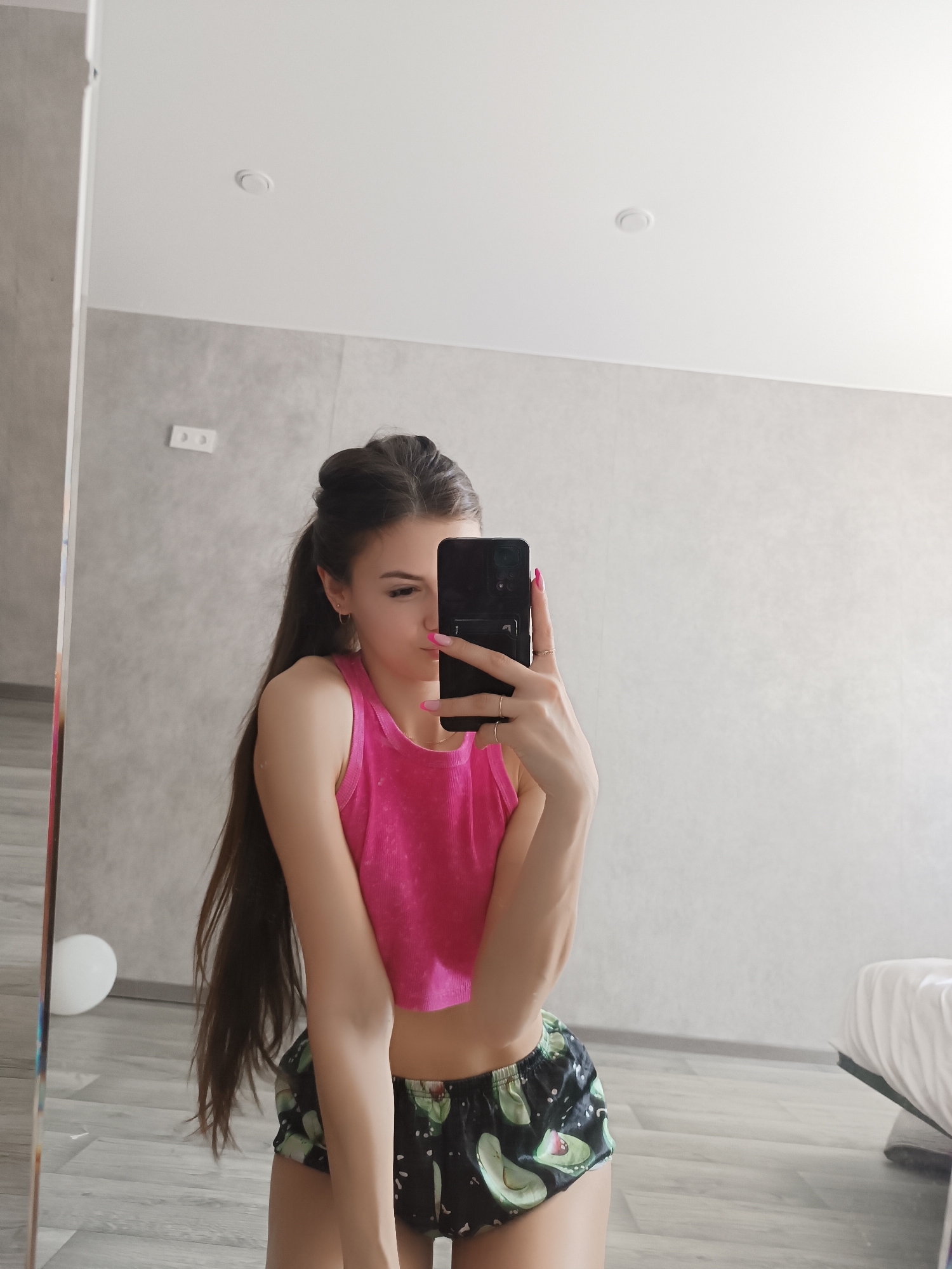 A woman stands indoors, holding a black smartphone in her right hand, presumably taking a selfie in a mirror. She is wearing a pink crop top and avocado-patterned shorts. Her long, dark hair is partially pulled back. The background includes a light gray wall with electrical outlets, a white object (possibly a balloon or a lamp) on the left, and the edge of a bed on the right. The location is potentially Chaikovsky, Russia, based on assumptions.