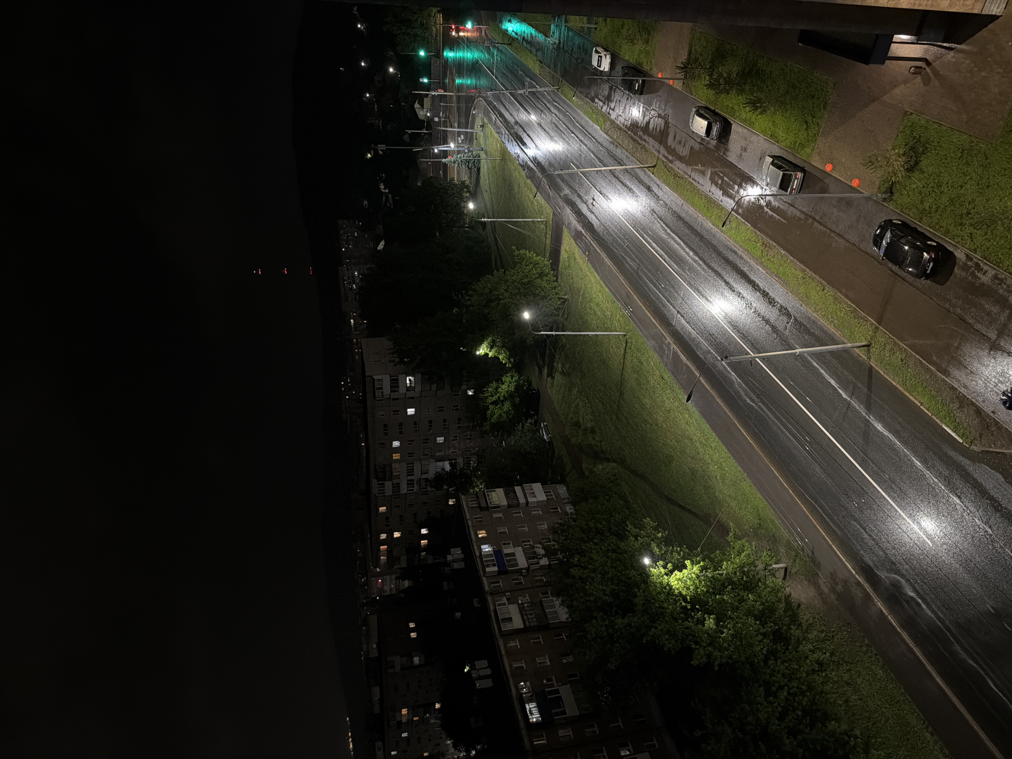 The image shows an aerial view of a wet urban street at night. The street is lined with greenery and streetlights, with a few cars parked on the side. In the background, buildings with illuminated windows can be seen. The lighting is dim, with reflections off the wet road surface and highlights from the streetlights and car headlights. The dark area on the left suggests a nighttime environment in the city of Unknown, Unknown.