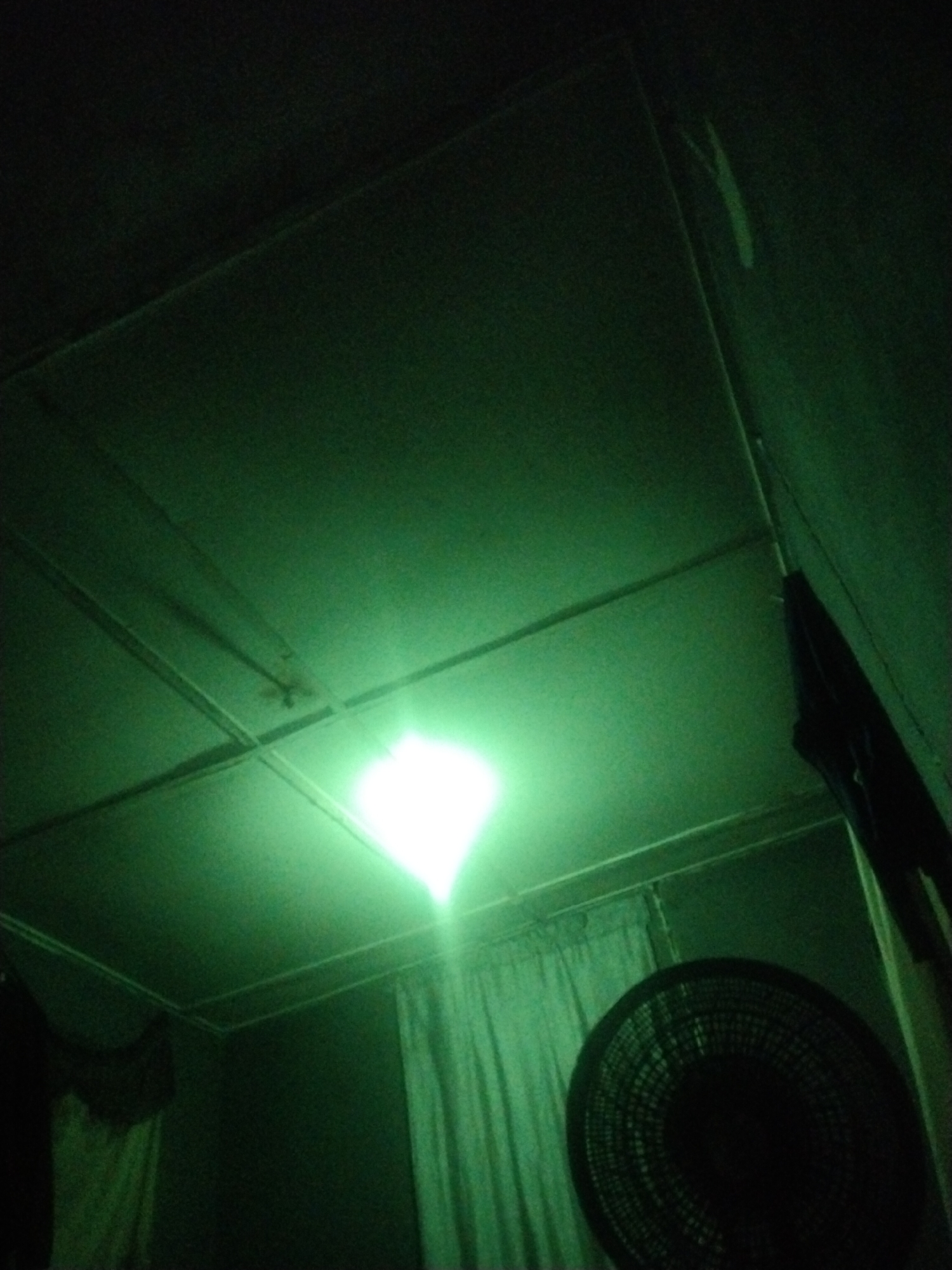 The image shows an interior view of a room, likely in Uyo, Nigeria. The illumination is green, likely from an LED light fixture on the ceiling, consisting of modular panels with visible seams. The ceiling appears low. A dark, circular electric fan is visible in the lower right corner of the frame. Curtains hang near the fan. Dark garments hang on the wall in the right side of the image.