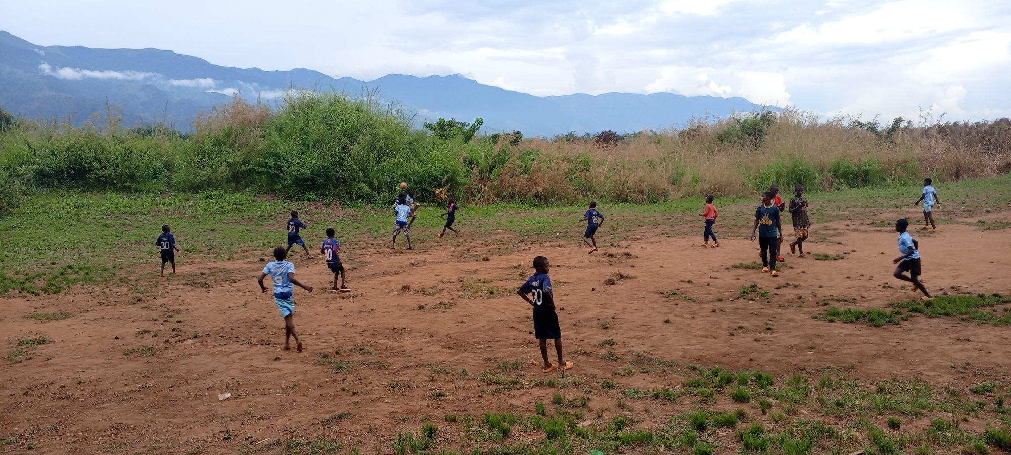 The image depicts an energetic outdoor scene of approximately 15-20 children, mostly boys, playing an informal game of football in what appears to be a rural or semi-rural setting. The location is Ngalula, Democratic Republic of the Congo.
The children are spread across a wide, open field with reddish-brown dirt, sparsely covered by patches of green grass. In the center of the activity, a light brown football is suspended in mid-air, indicating an active moment of play, likely a header or a kick. Several children are gathered around the ball, actively engaged, while others are positioned across the field, running or observing. Most of the children are barefoot or wearing simple sandals.
Many children are wearing football jerseys. Visible text includes "MESSI 30" on at least three blue jerseys, "MESSI 20" on another, and "JOHN 10" on a light blue and pink jersey. One child wears a dark t-shirt featuring a white "JEEP" logo.
The background is dominated by lush, dense green vegetation, including tall grasses and bushes, forming a natural boundary for the playing area. Beyond this greenery, a majestic mountain range rises, its peaks partially obscured by soft white and grey clouds, suggesting an overcast sky. The lighting is diffused and soft, indicating daytime, possibly an overcast morning or late afternoon. The overall atmosphere conveys a sense of active play and community enjoyment amidst a natural, picturesque landscape.