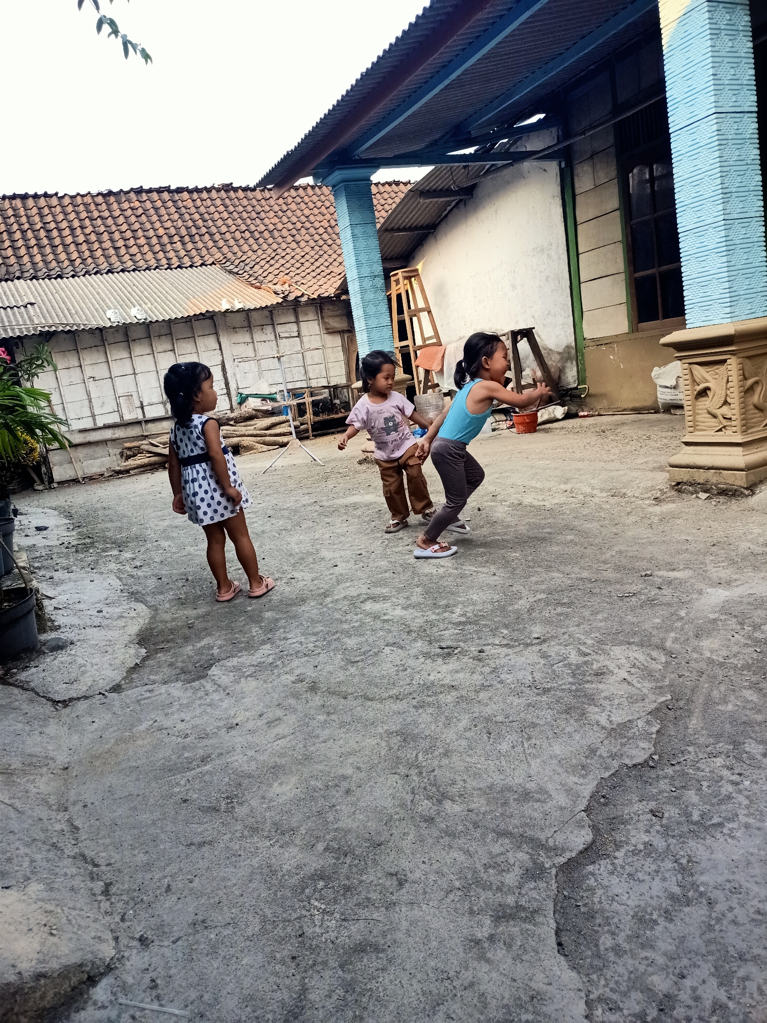 In Kendung, Indonesia, three young girls are playing in a concrete courtyard. The girl on the left wears a white dress with blue polka dots and pink sandals. The girl in the middle has brown hair and wears a pink t-shirt and brown pants. The girl on the right, with her hair in a ponytail, is dressed in a blue tank top and dark leggings, with white sandals. She appears to be in motion, reaching out her right arm. The background features a building with a corrugated metal roof, white siding, and a light blue decorative pillar. The ground is a rough, grey concrete surface with some cracks and debris.