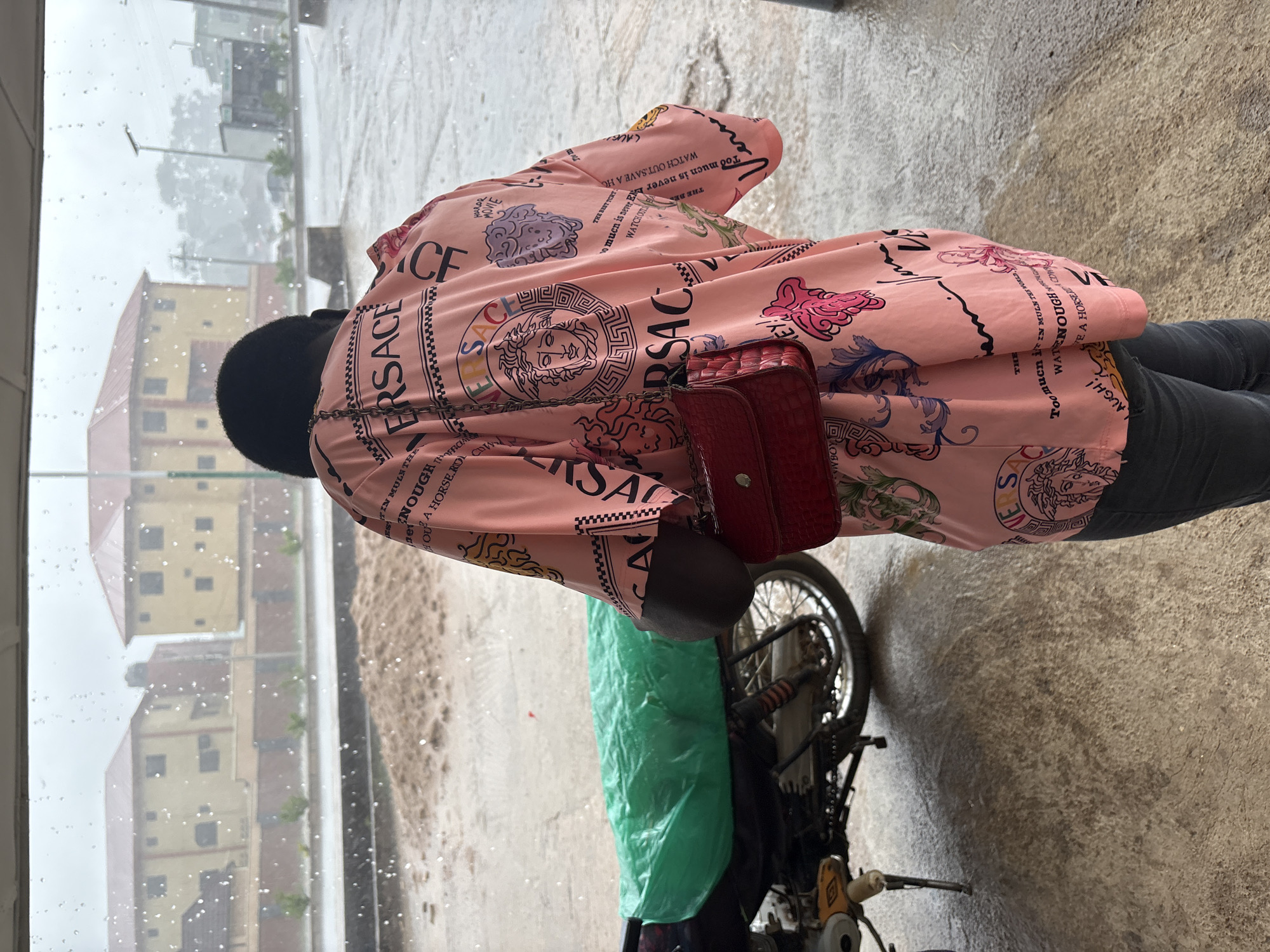 A person in a pink Versace shirt and dark pants stands next to a motorcycle, while holding a small red purse. The motorcycle is partially covered with green plastic. Rain is visible on the window, obscuring a view of the city, potentially Bukuru, Nigeria. Buildings can be seen in the background. The surface is concrete and damp.
