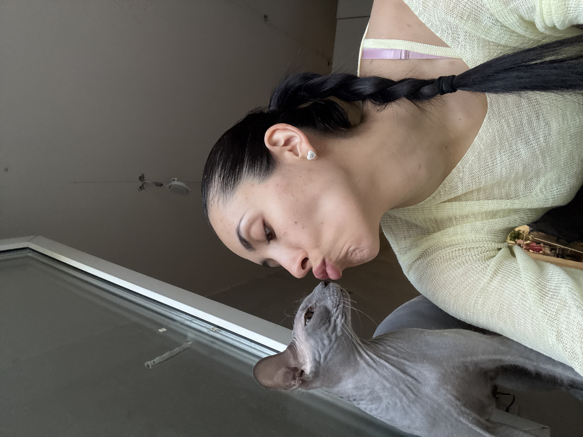 An indoor scene depicts a woman and a grey Sphynx cat. The woman, shown in profile, has dark hair styled in a tight, low braid resting over her right shoulder. She wears a light-colored, loose-knit v-neck top, with a pink bra strap visible on her right shoulder. A white heart-shaped stud earring is present in her right ear. Her lips are pursed, directed towards the cat's face. The Sphynx cat, positioned adjacent to a window with a white frame, faces the woman with its head slightly turned away from the camera. The background consists of a dark windowpane behind the cat, a light-toned ceiling displaying a partially visible white circular fixture suspended by a wire, and a muted wall surface to the right. The scene is located in the city of Votkinsk, Russia.