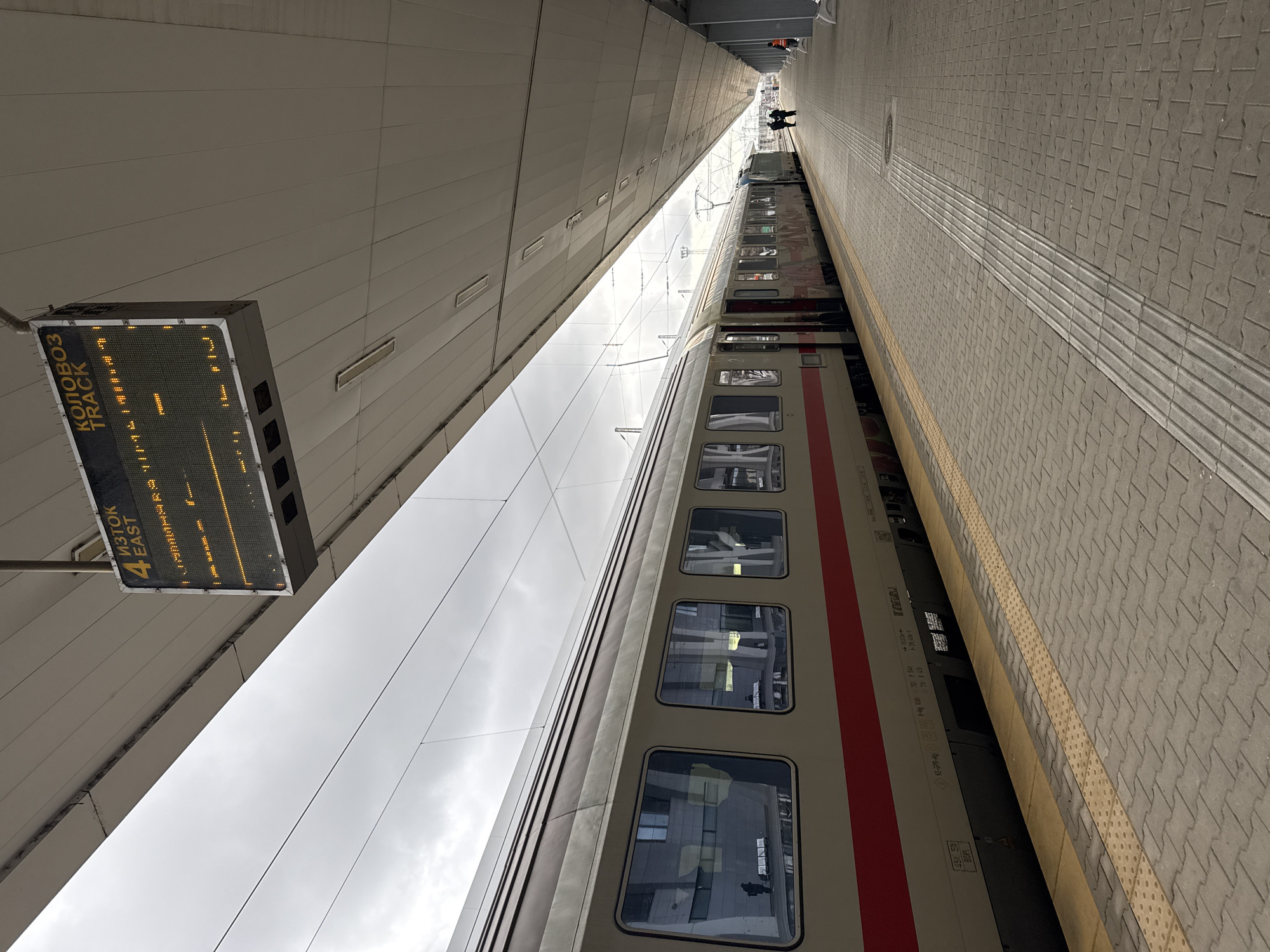 This image depicts a train at a station in Sofia, Bulgaria. The train is beige with a red stripe. The station has a gray concrete platform with a tactile strip for the visually impaired. Above the train, there is a sign displaying the track number as "4 EAST" and the destination in Cyrillic, "КОЛОВОЗ" (Track). People are visible in the distance at the end of the platform. The station's architecture is modern with a high ceiling and reflective paneling. The sky is overcast.