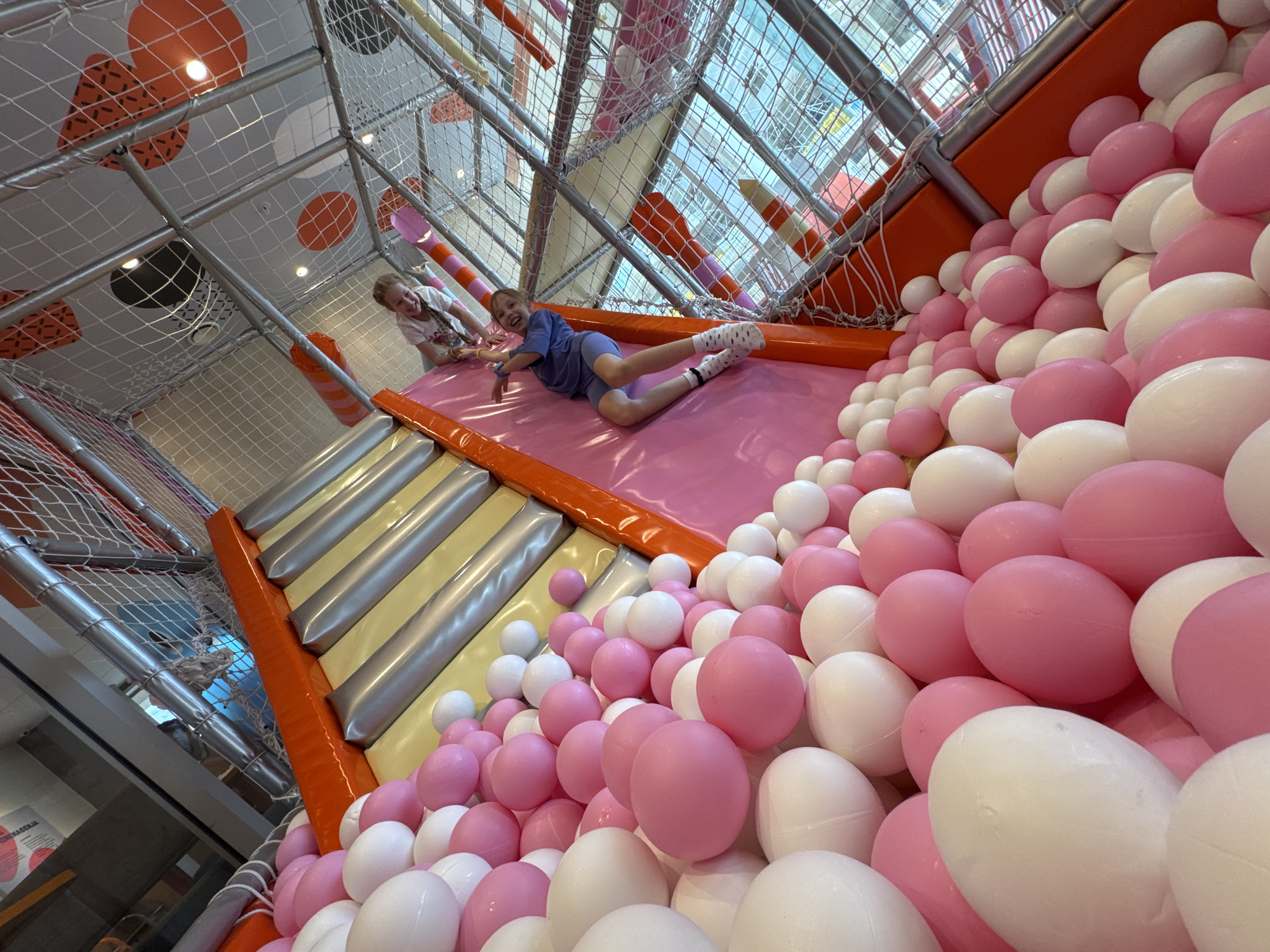 Two girls are playing in an indoor playground. One girl is lying on her stomach on a pink slide, while the other girl stands at the top of the slide. A large pile of pink and white balls is adjacent to the slide. The playground appears to be indoors, with a metal frame structure and netting. The location appears to be in Budva, Montenegro.