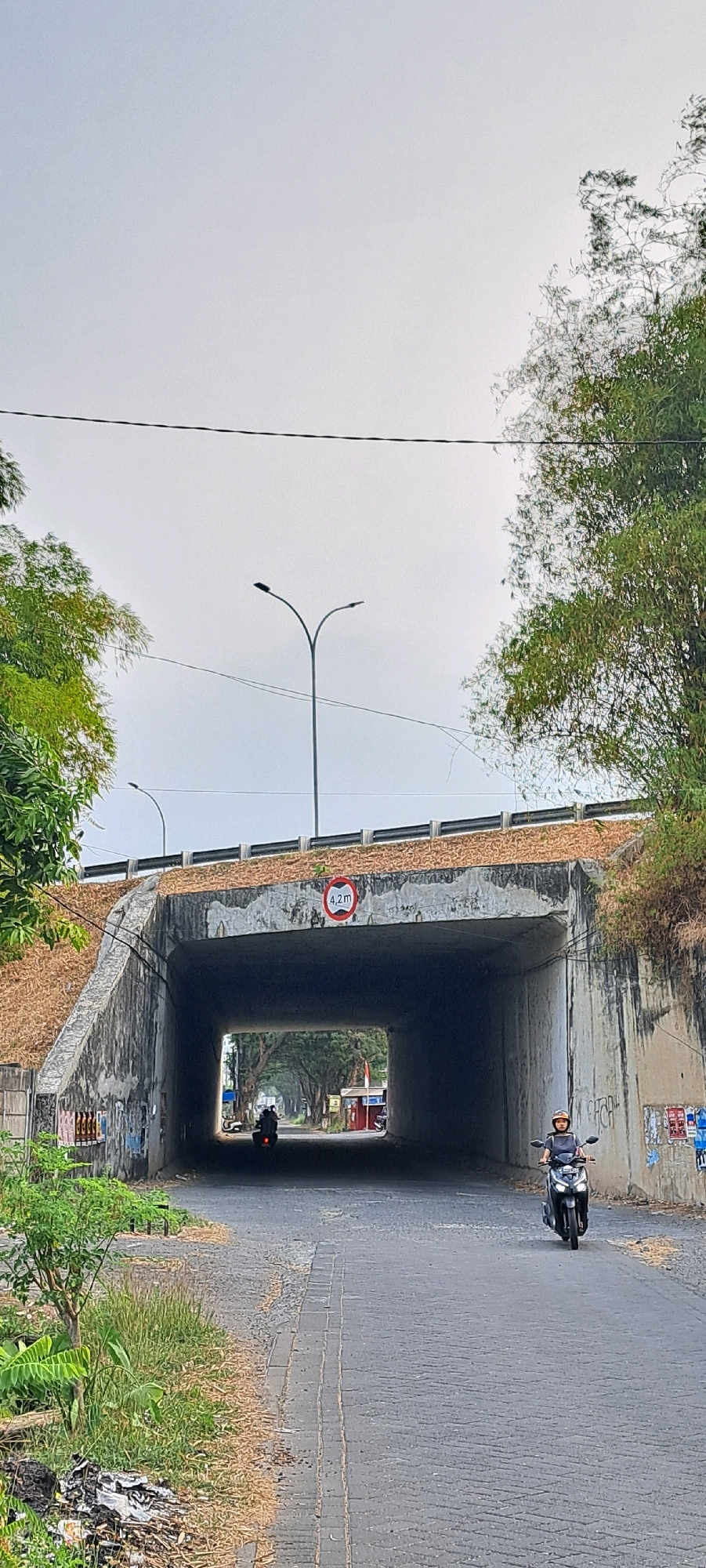 This is a vertical, eye-level shot of a concrete underpass on a road in Wedoro, Indonesia, on an overcast day. The underpass is lined with concrete on the left and right, and the tunnel opening reveals trees and buildings in the distance. A sign indicating "4.2 m" in red is affixed to the right side of the underpass's exterior. A paved road with a textured surface leads into the tunnel. On the right side of the road, a person wearing a helmet rides a black motorcycle into the tunnel. On the left side of the road, another motorcycle is partially visible entering the tunnel. The grassy banks on either side of the road are covered with dry leaves and debris. Green foliage and trees are visible on the slopes above the underpass. Power lines are strung across the sky.