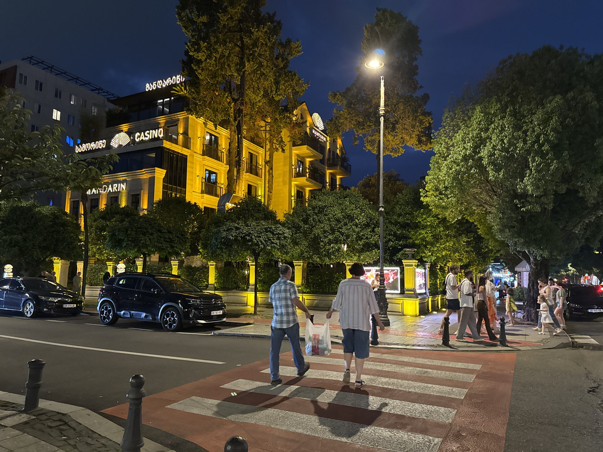 The image shows a street scene at night in Batumi, Georgia. A building with "MANDARIN CASINO" signage is illuminated by yellow lights. Several people are crossing a pedestrian walkway on the street. Two individuals in the foreground are walking across the street carrying a grocery bag. A black SUV and a dark sedan are parked on the side of the street. The sky is dark blue, indicating nighttime. Lush green trees line the street, and a streetlamp illuminates the area. The building appears to be a hotel or a resort.
