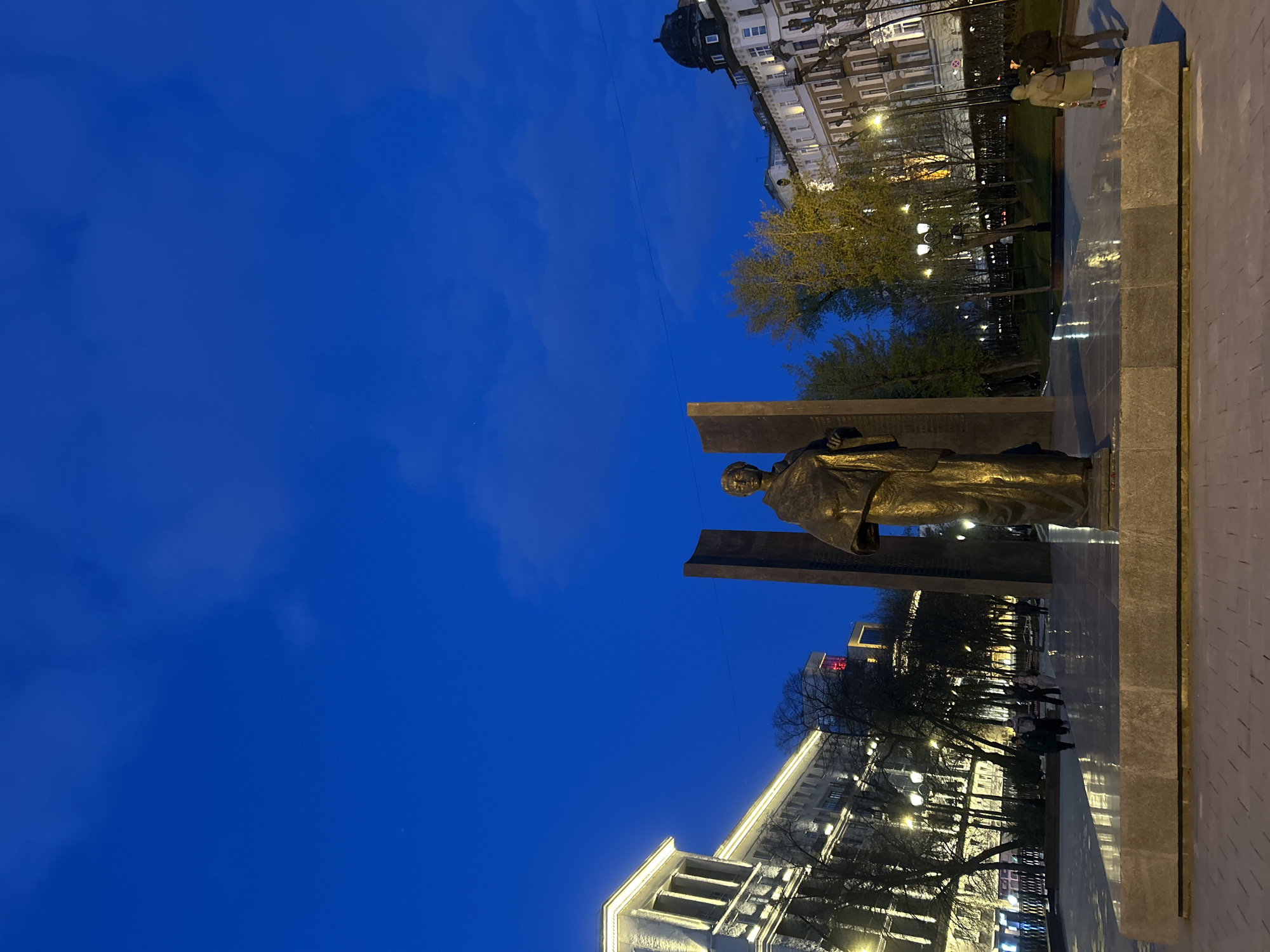 The image, oriented vertically, captures a city scene at dusk or early night, characterized by a deep blue sky with subtle cloud formations. In the foreground, a bronze statue of a standing figure, likely a historical personage, is positioned on a substantial polished stone plinth. The statue is flanked by large, rectangular stone steles.
To the left of the monument, a row of impressive, classically styled buildings with illuminated facades stretches into the background. Bare trees and others showing early signs of foliage line the paved pathways, interspersed with numerous glowing streetlights that cast warm light on the surroundings.
On the right side of the frame, more grand architectural structures are visible against the twilight sky, including one building topped with a dark dome. Several pedestrians can be seen walking on the paved areas, with one individual in a light-colored coat particularly noticeable near the base of the monument. The artificial lighting from the buildings and streetlamps creates a serene ambiance, reflecting off the smooth surfaces of the pavement. The setting, with its distinctive architecture and monument, is typical of a large Russian city like Moscow. No legible text is visible on the monument or buildings.