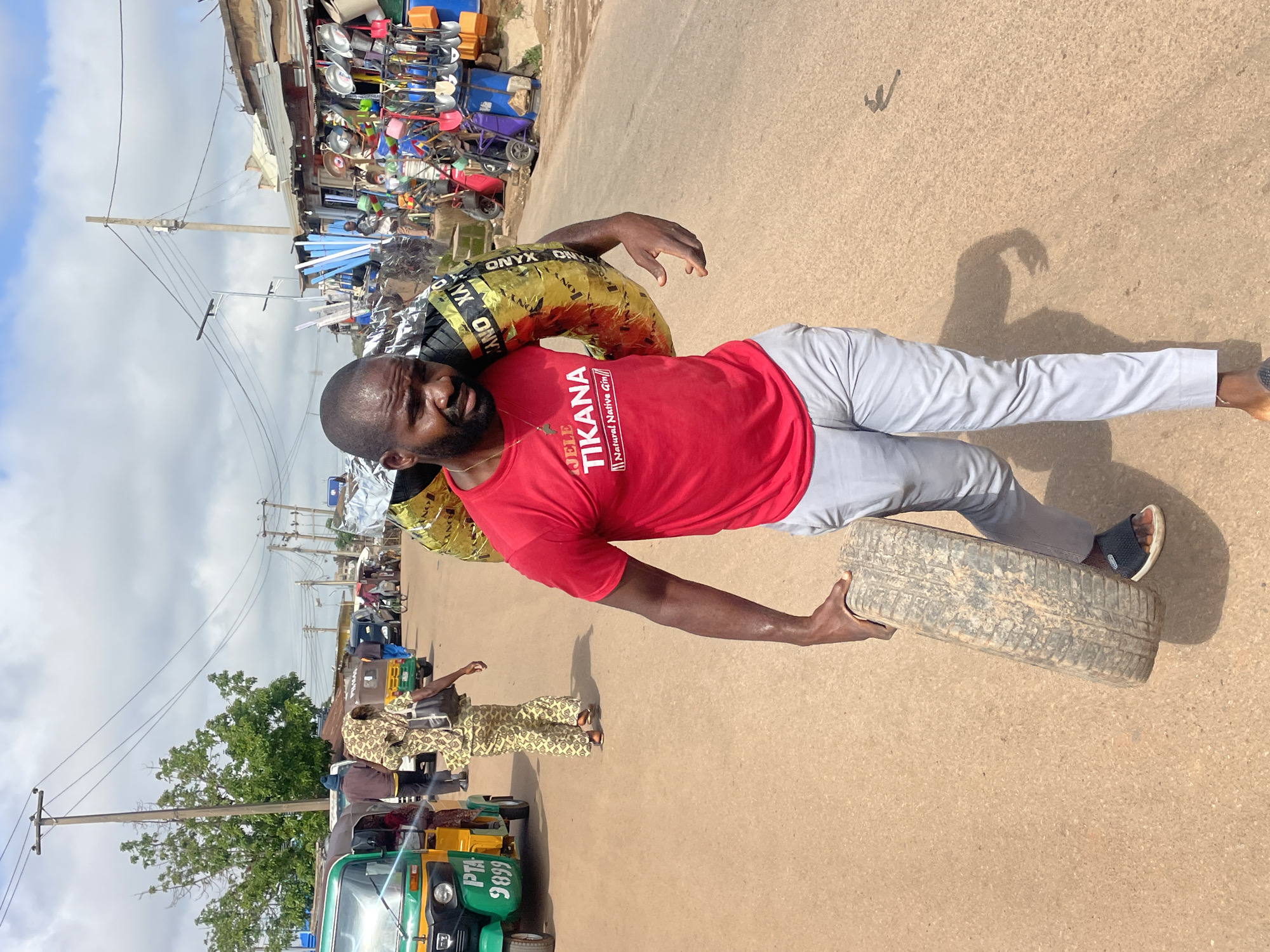 In Gura Top, Nigeria, a man in a red t-shirt with "JELE TIKANA Natural Native Gin" printed on it, and light grey trousers, walks down a dusty street carrying a large tire on his right shoulder, another tire in his right hand, and a third tire wrapped in golden material over his left shoulder. He is wearing dark sandals. Behind him, a woman in a patterned outfit walks away from the camera, carrying a dark bag. Several vehicles, including a green and yellow auto-rickshaw with "PTA 9899" on it, are visible on the street, along with market stalls displaying various goods such as tools and household items. The sky is cloudy, and utility poles with power lines are present.