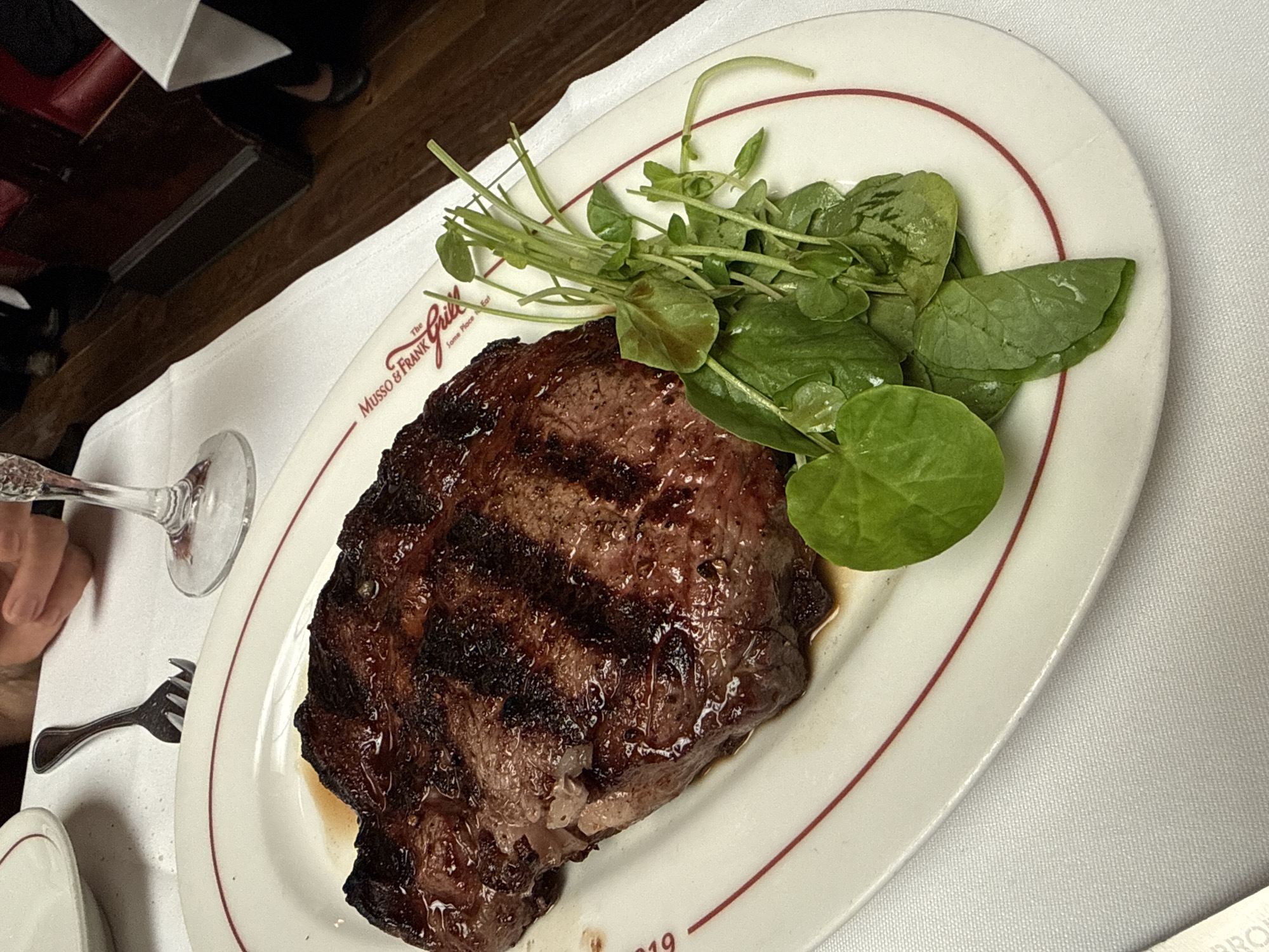 A close-up, overhead shot features a thick, juicy steak grilled to perfection, served on a white oval plate with a red rim. The steak is glossy and has prominent grill marks. To the right of the steak, a small pile of fresh, green watercress garnish is arranged. The plate rests on a white tablecloth. In the background, a hand holds a fork, and a wine glass is visible on the table. Part of a restaurant interior, with dark wood paneling and red seating, is also discernible. The plate bears the embossed logo "MUSSO & FRANK GRILL" which is a historic Hollywood restaurant located at 6667 Hollywood Boulevard, Los Angeles, California, United States of America.
