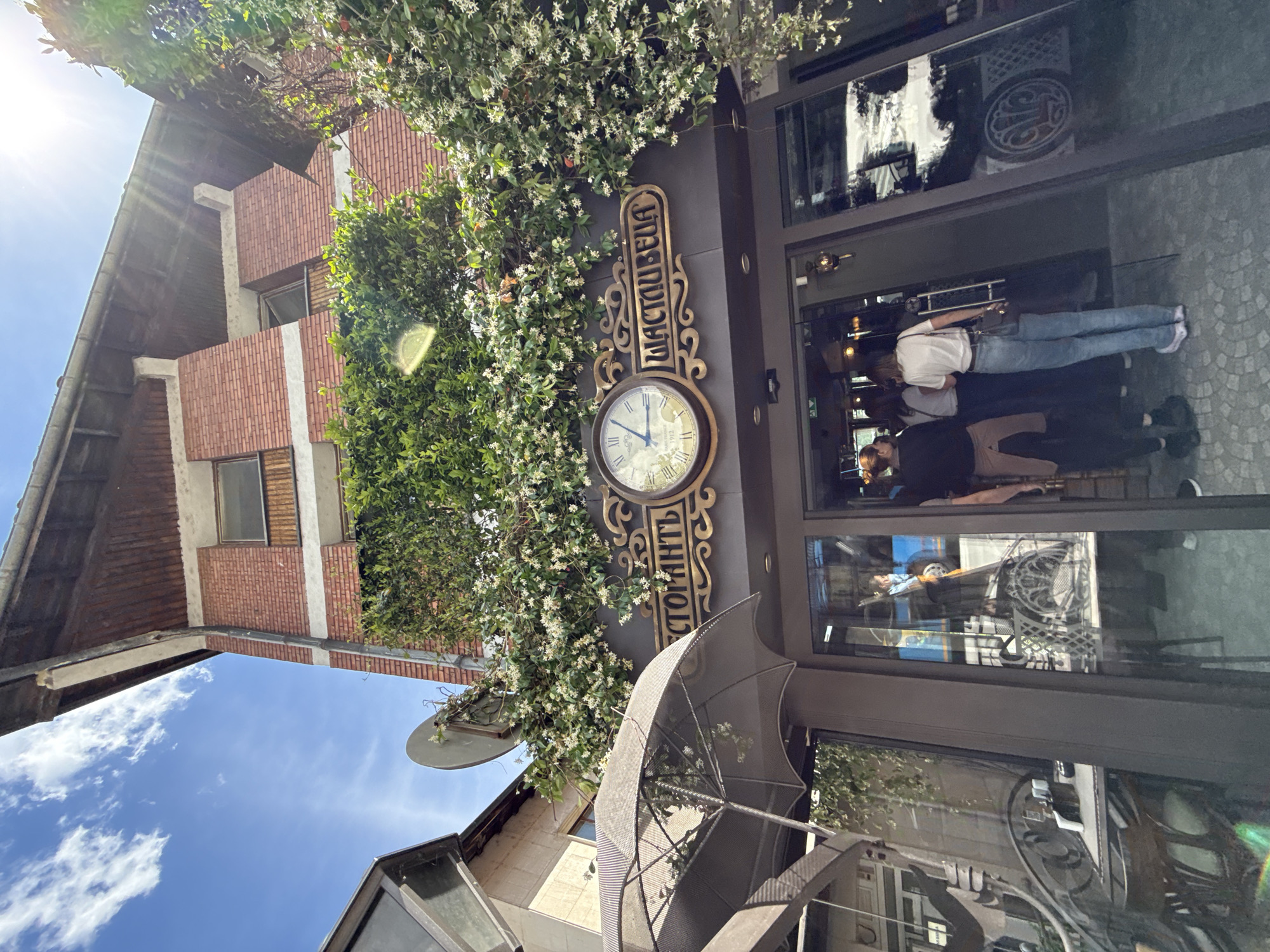 The image shows the exterior of a restaurant named "Tsastvelella" in Veliko Tarnovo, Bulgaria. The restaurant's entrance is adorned with greenery and a decorative clock. Several people are visible inside the restaurant, reflected in the glass entrance. An umbrella stand is positioned outside the entrance. The architecture surrounding the restaurant includes a brick building and other structures under a blue sky.