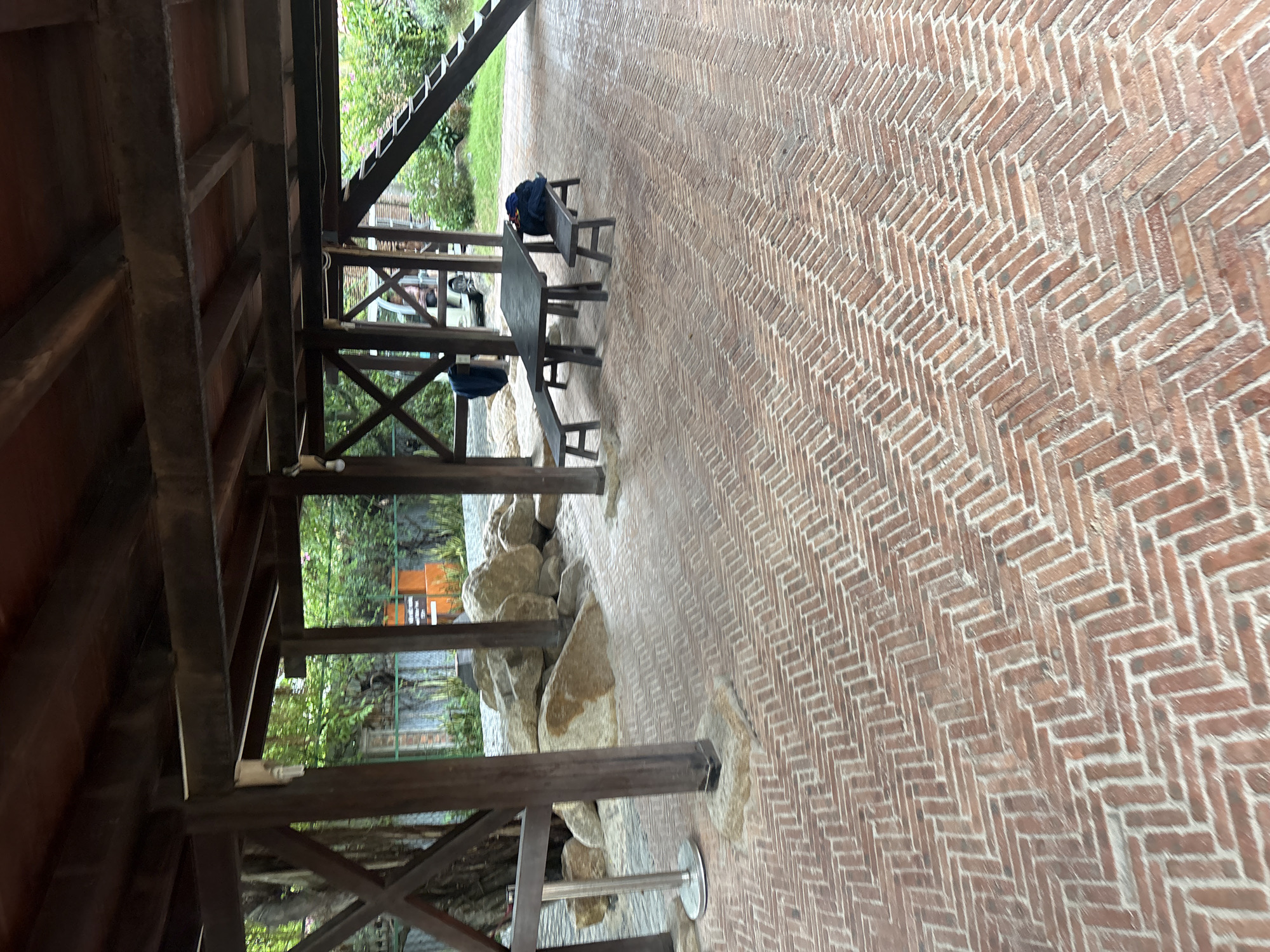 The image shows a wooden structure with a red brick herringbone-patterned floor. Underneath the structure, a dark rectangular table with two dark benches is positioned. Bags are placed on the benches. The area surrounding the structure features large rocks and lush greenery. In the background, a white vehicle and a set of stairs leading upwards are visible. The scene appears to be an outdoor seating area, possibly a part of a park or a recreational space. The location is identified as Nha Trang, Vietnam.