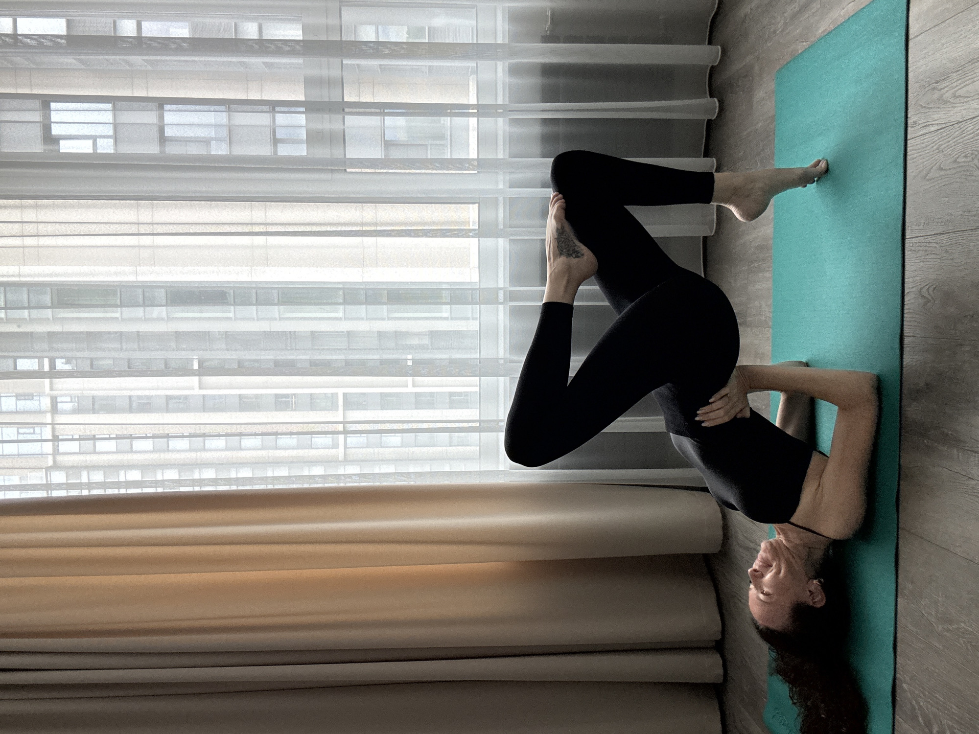 A woman in a black athletic outfit is performing a yoga pose on a teal mat placed on a wooden floor. Her body is arched upwards, with her hands clasped behind her head, supporting her upper back. Her legs are bent, with one foot flat on the mat and the other crossed over the bent knee, revealing a tattoo on her ankle. The woman's head is tilted back, and she appears to be smiling or grimacing. The background features sheer white curtains covering a large window, through which the facade of a modern building with numerous windows is visible. A beige curtain is drawn to the left side of the window. This scene is indoors, likely in an apartment or studio, given the domestic setting and the view of a city building. The location is specified as being in the city of Khimki, Russia.