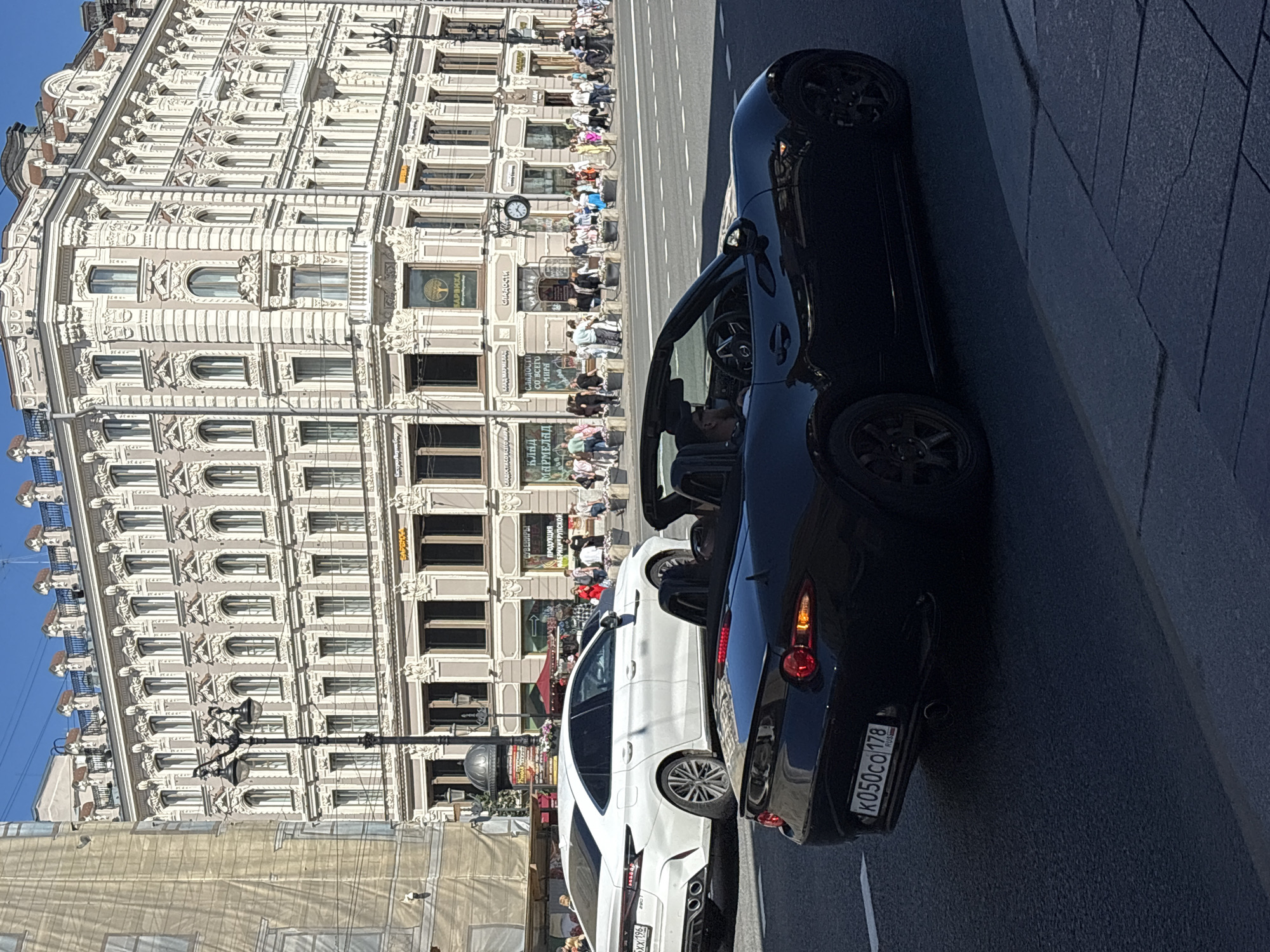 The image captures a sunny street scene in Saint Petersburg, Russia, with a focus on architectural detail and vehicular traffic. A light-colored, modern sedan is parked on the left side of the street, its rear visible. In the foreground, a black convertible is also on the street, partially obscuring the sedan. The car's interior shows a driver wearing sunglasses and a cap.
The backdrop is dominated by a grand, ornate building with multiple stories, showcasing intricate classical architectural elements such as columns, decorative moldings, and arched windows. The building appears to be a commercial establishment, with signage visible, including what appears to be "СЛАДОСТИ" (sweets) and "КЛАД МАРМЕЛАД" (marmalade treasure).
A sidewalk in front of the building is lined with people, suggesting a bustling urban environment. Some individuals are walking, while others appear to be congregating. The street is paved, and road markings are visible, indicating it is an active thoroughfare. A lamppost with multiple lights stands near the sidewalk. The sky is clear and blue, indicating fair weather.