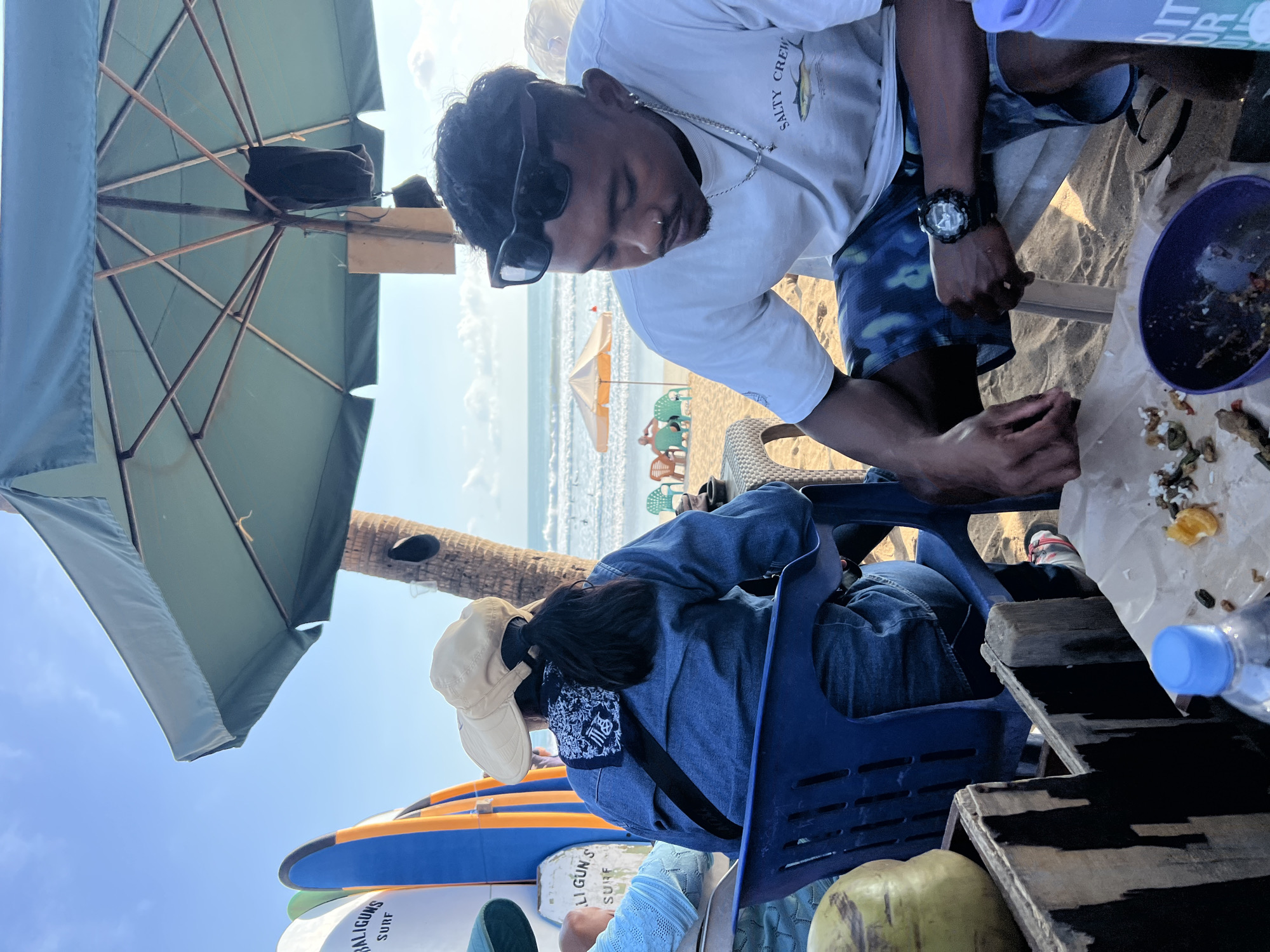 This is a medium shot of two individuals seated at a table on a beach in Kuta, Indonesia, under a large, open umbrella. The person on the right, a male, is in the foreground, looking down at food on the table. He is wearing a white t-shirt with the words "SALTY CREW" and a fish graphic, and blue patterned shorts. He has sunglasses perched on his head, a necklace, and a watch on his left wrist. The person on the left, facing away from the camera, is wearing a denim jacket, a beige hat, and a bandana around their neck. Surfboards are visible in the background to the left. The beach and ocean are visible in the distance, with a few people seated at tables further down the shore. The lighting suggests it is late afternoon.