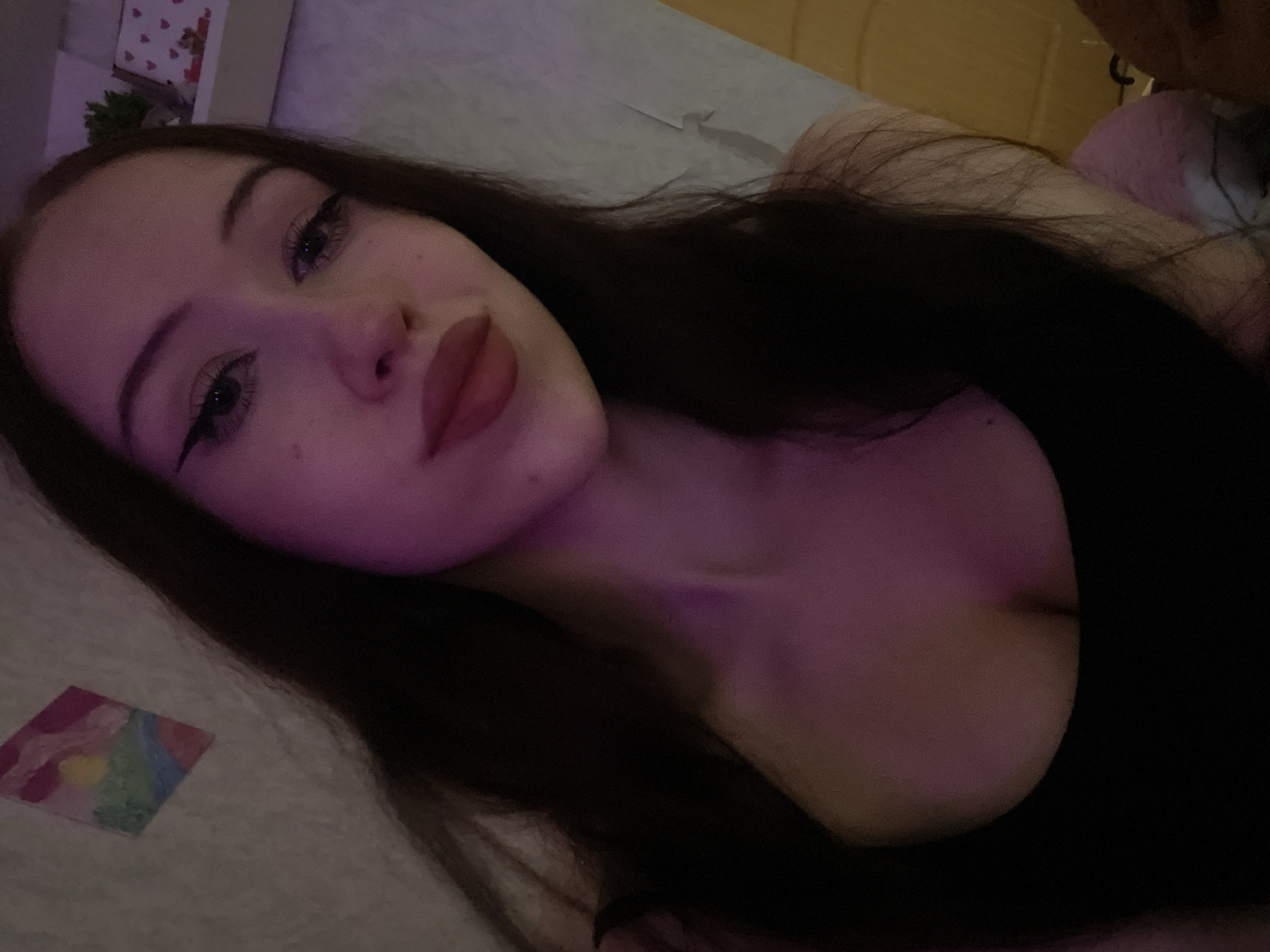 A selfie taken indoors, likely in a residential setting in Votkinsk, Russia, features a young woman with long, dark hair. She is looking directly at the camera, with her head slightly tilted. Her makeup includes winged eyeliner and matte lipstick in a reddish-brown shade. She is wearing a black sleeveless top that reveals part of her chest. The background is softly lit with purple and pink hues, and a small, colorful painting can be seen on the wall to her left. To her right, a shelf with some decorative items, including small heart-shaped figures and a small potted plant, is partially visible. The overall atmosphere of the image is intimate and personal.