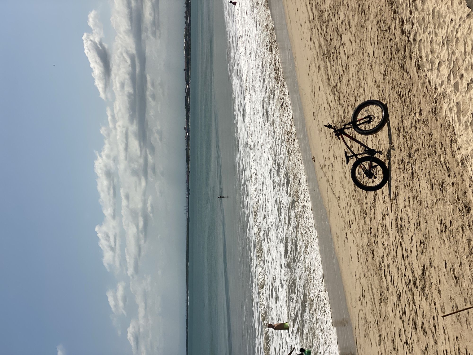 The image depicts a beach scene in Kuta Selatan, Indonesia. A bicycle is parked on the sandy shore. Waves are crashing on the beach. In the distance, a shoreline and city buildings are visible. The sky is partially cloudy. A few people are scattered along the beach and in the water.