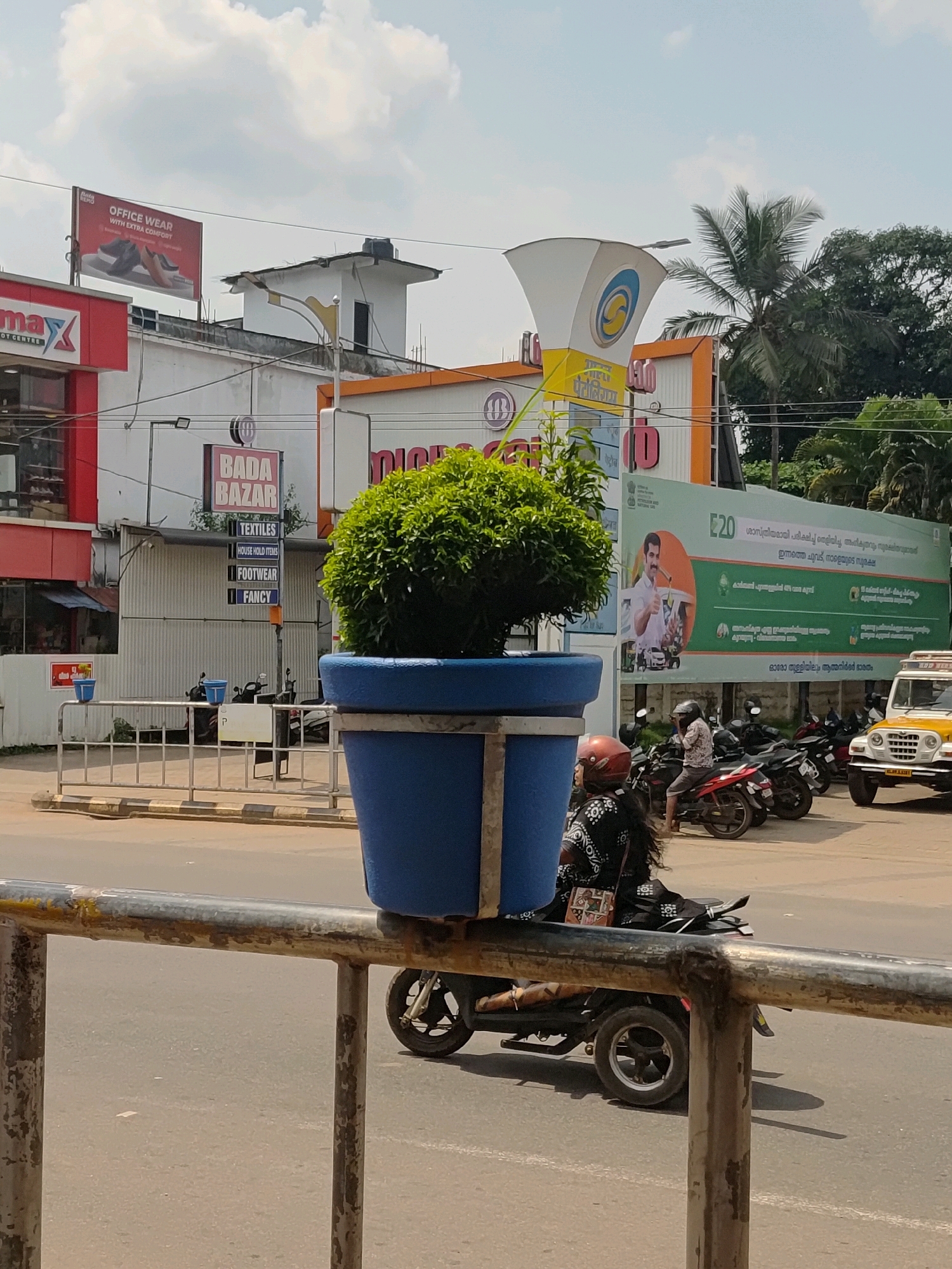 In Mannarkkad, India, a street scene unfolds under a bright, partly cloudy sky. A large, blue planter pot with a lush green shrub sits prominently in the foreground. Behind it, a metal railing divides the foreground from the street. The street itself is busy with motorcycles and a yellow jeep.
On the left side of the image, a red building with "max" written on it is visible. Above this, a billboard advertising "OFFICE WEAR WITH EXTRA COMFORT" is mounted. To the right of this building, another structure features a sign that reads "BADA BAZAR" with sub-sections for "TEXTILES," "HOUSE HOLD ITEMS," "FOOTWEAR," and "FANCY." Further to the right, a large green billboard displays an image of a man giving a thumbs-up, accompanied by text in Malayalam. A tall, conical structure with a circular logo is also visible, likely part of a business establishment. The background consists of trees and buildings, indicating an urban or semi-urban setting. A woman wearing a black and white patterned dress rides a scooter, and another person on a scooter is partially visible. The overall impression is of a commercial street with active transportation.