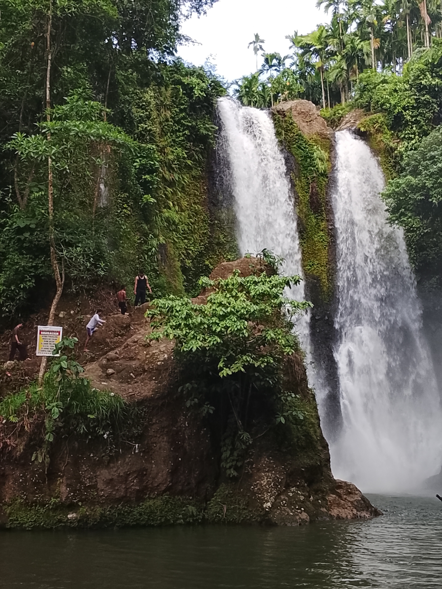 A wide shot captures a cascading waterfall in a lush, tropical environment. The waterfall is split into multiple streams, with water plunging over a rocky cliff face covered in green moss and vegetation. In the foreground, dark, calm water reflects the greenery. On the rocky bank to the left of the waterfall, a path is visible, with several people making their way up or standing near it. One individual, wearing a black tank top and dark shorts, stands prominently on a higher section of the path. Another person in a white t-shirt and blue shorts is seen climbing. Further down the path, two more individuals are present. A signpost with some text is also visible near the path. The top of the image is framed by dense green foliage and tall palm trees. The location is believed to be the Jantong Hujan waterfall in Lhokseumawe, Indonesia.