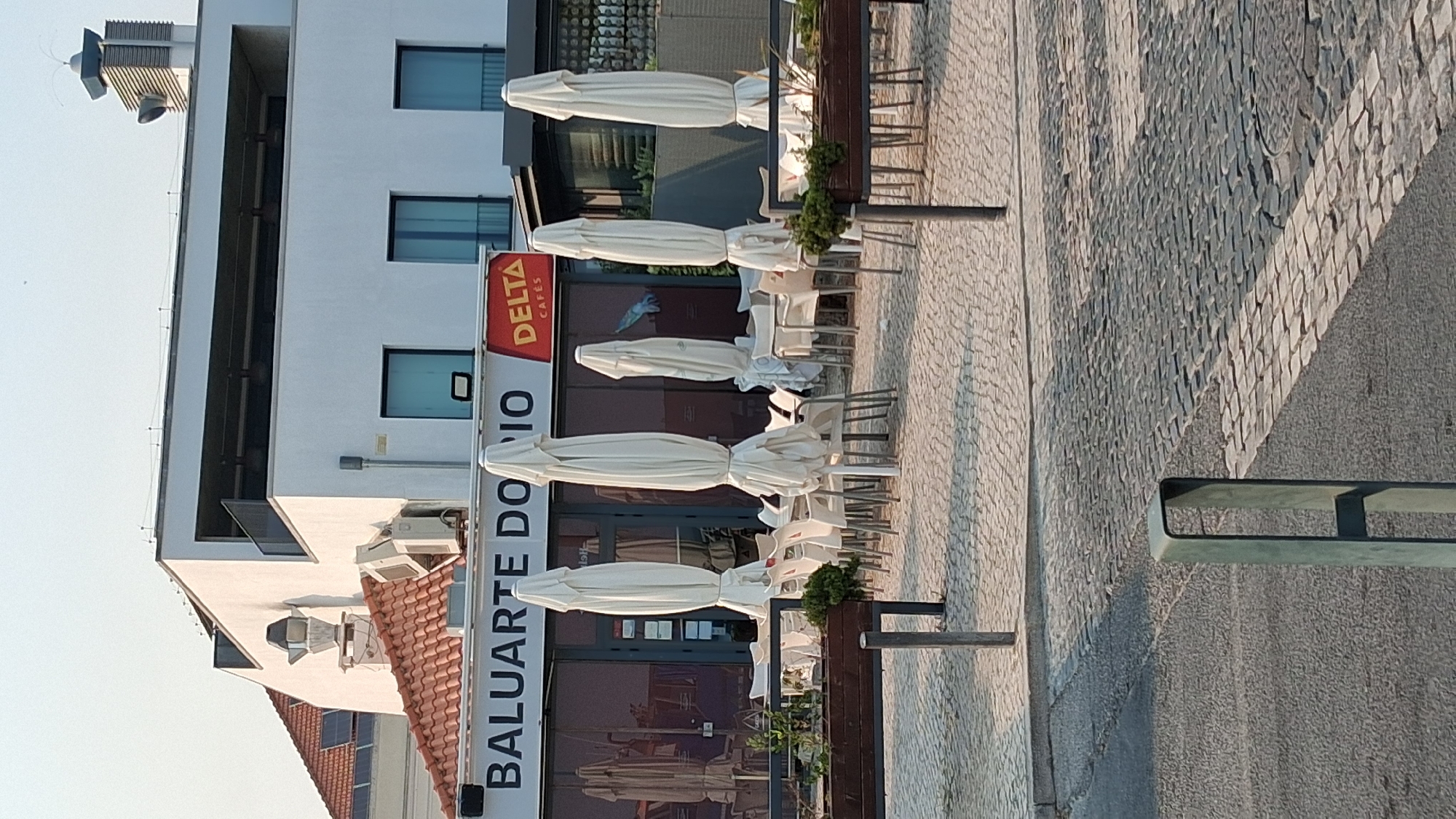 The image depicts the exterior of a café, identified by the signage "BALUARTE DO RIO DELTA CAFÉS". The café appears to be located in Setúbal, Portugal, near the waterfront, possibly along the Avenida Luísa Todi, given the "RIO" in the name and the style of building and paving. The scene is set during daylight, with the sun casting shadows. In the foreground, there is a cobblestone pavement and a dark grey road. Several white, closed patio umbrellas are prominent, arranged around tables and chairs that are set up outside the establishment. Some of these tables and chairs are empty. The building behind the café seating area is modern with white walls and large windows. There are no people visible in the immediate vicinity of the tables and chairs. The overall impression is of an outdoor seating area of a closed or preparing-to-open café.