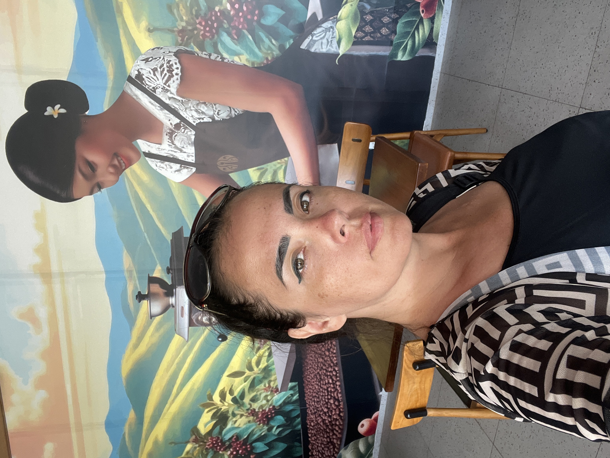 This is a portrait-style photograph taken with a shallow depth of field, focusing on a woman in the foreground. She is looking directly at the camera, with a neutral to slightly inquisitive expression. She has dark hair pulled back, sunglasses perched on her head, and is wearing a black top with a patterned overlay.
Behind her is a mural depicting a coffee harvesting scene. The mural features a woman in traditional attire with her hair in a bun, a coffee grinder, and a landscape of coffee plants with red berries. The style of the mural suggests a tropical or agricultural setting.
The overall lighting appears to be natural daylight, possibly indicating daytime. The background of the mural is vibrant, with blues, greens, and earthy tones, suggesting a sunny climate. The presence of wooden chairs and a stone-like wall suggests an indoor or semi-outdoor setting, possibly a cafe or restaurant in Kuta Selatan, Indonesia, given the location context. There is no visible text in the image. The image's orientation is vertical, with the woman's head at the top.