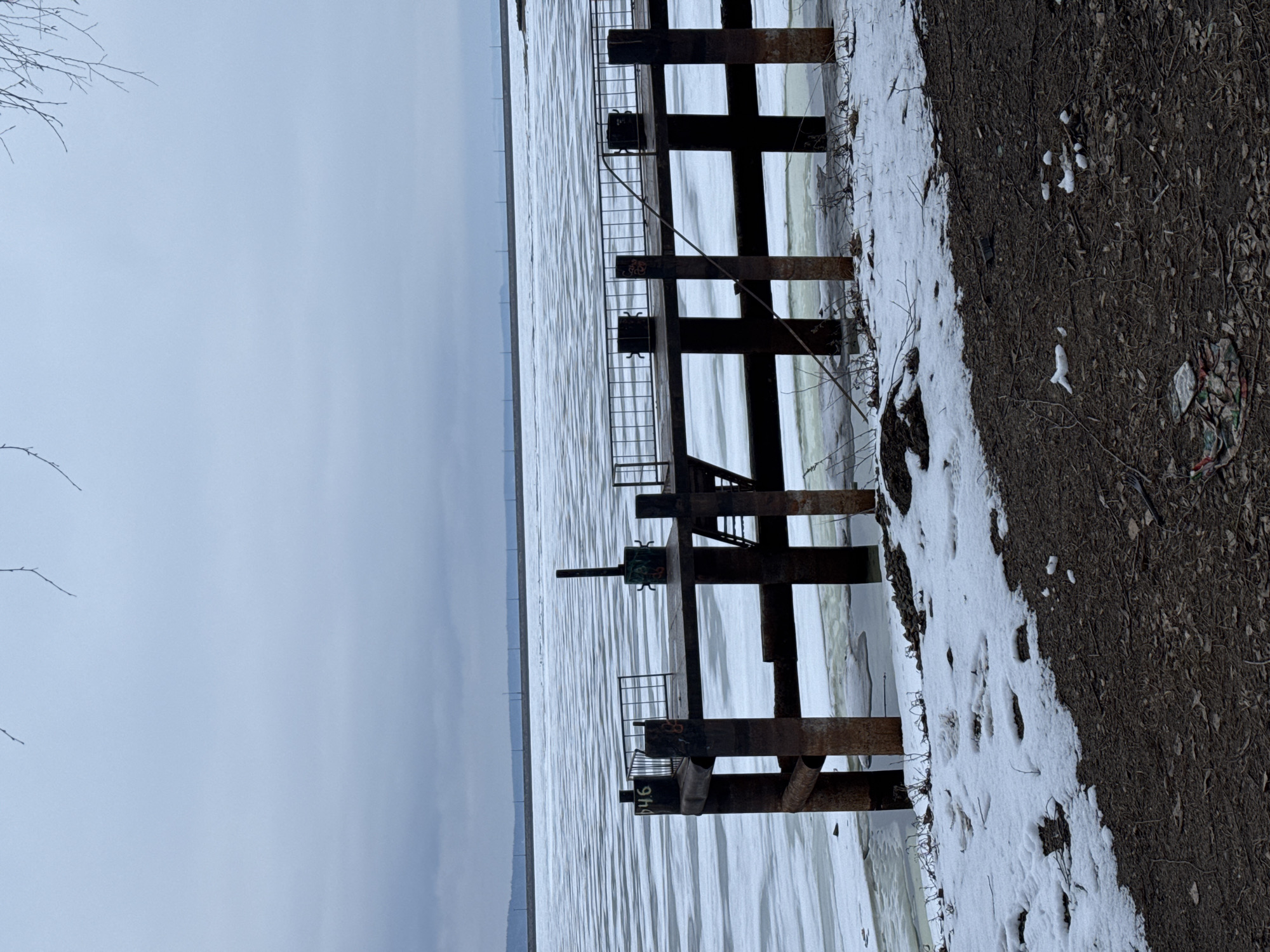 The image depicts a waterfront scene in Chaikovsky, Russia. A partially frozen body of water stretches into the distance under an overcast sky. A dark, rusted, metal pier-like structure with a ladder runs into the water. The shore is visible on the right side of the frame with snow cover near the structure and bare earth further back. Debris is scattered on the ground. No people are visible. The structure is marked with the number "646".