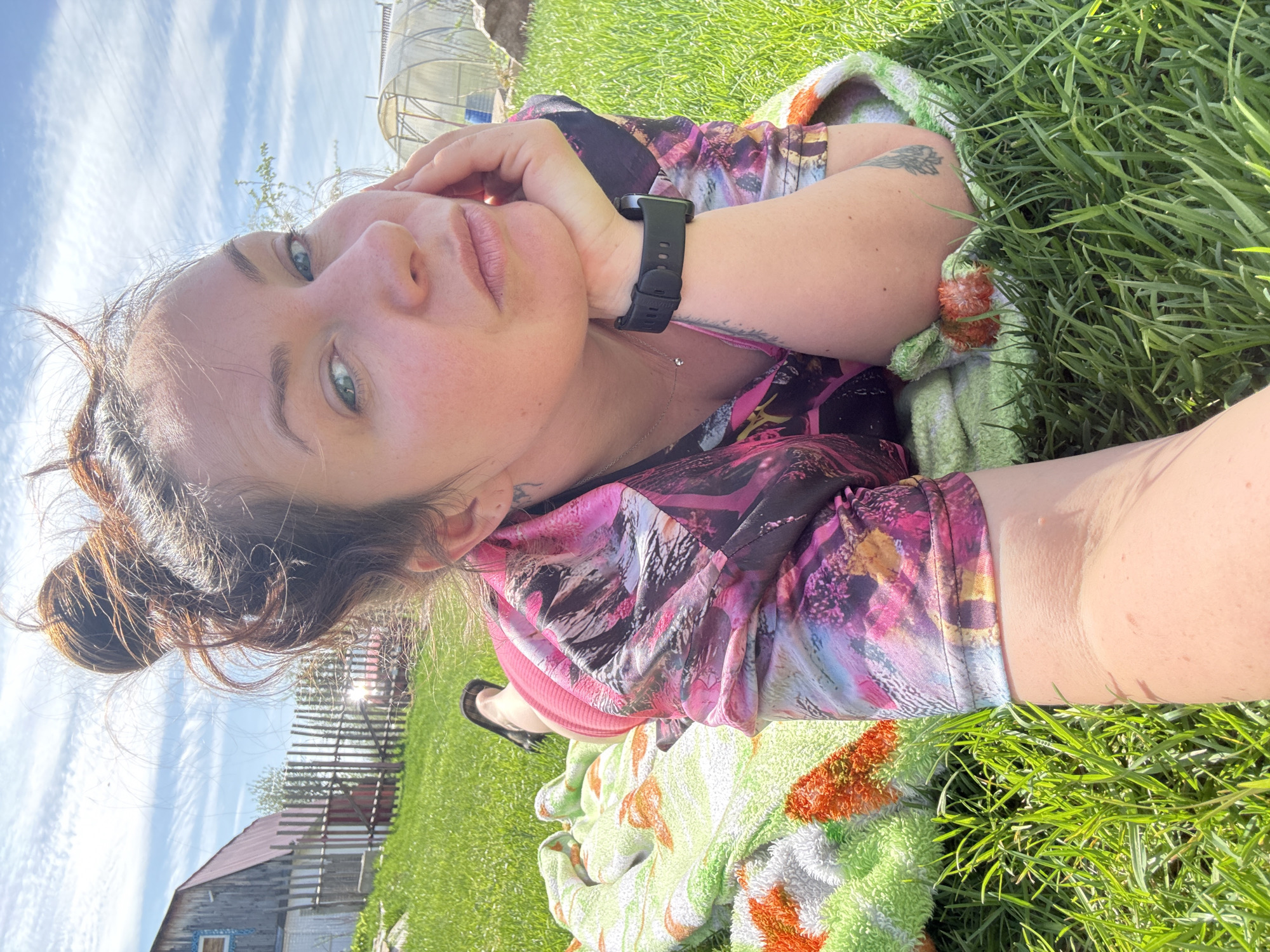 A woman lies on a green lawn, resting her head on her hand. She wears a multicolored top, a black smartwatch on her left wrist, and a thin necklace. She is partially covered by a light green towel with orange patterns. The background includes a wooden building, a metal fence, and a greenhouse. The sky is visible with wispy clouds. Based on the geolocation, the location is likely in Chaikovsky, Russia.